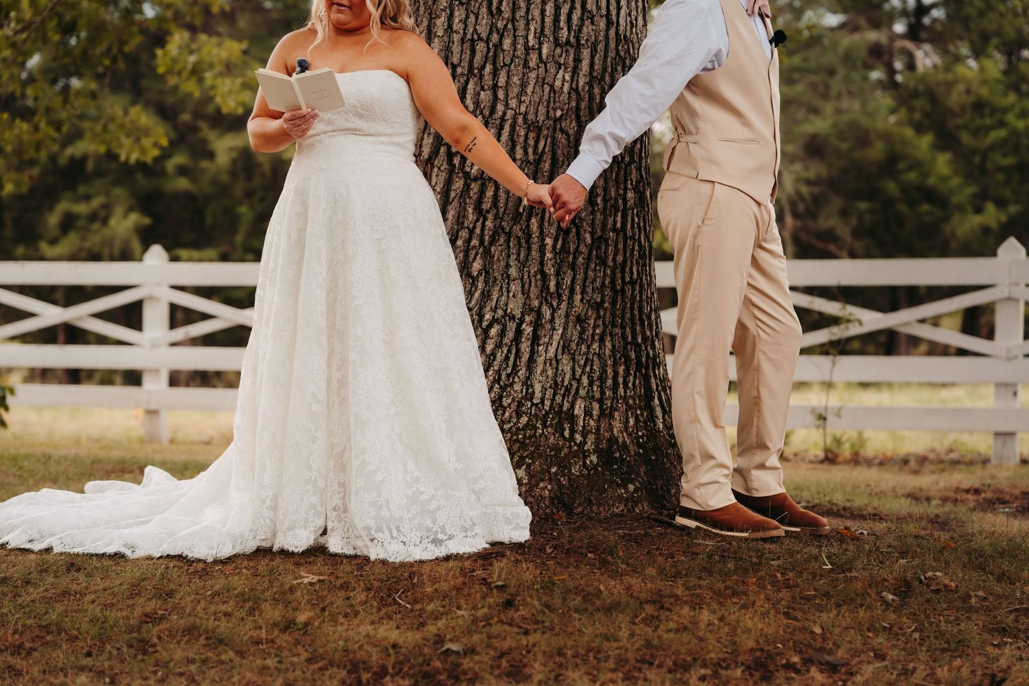 Bride and groom holding hands around a large tree trunk at an outdoor wedding ceremony with white fence backdrop.