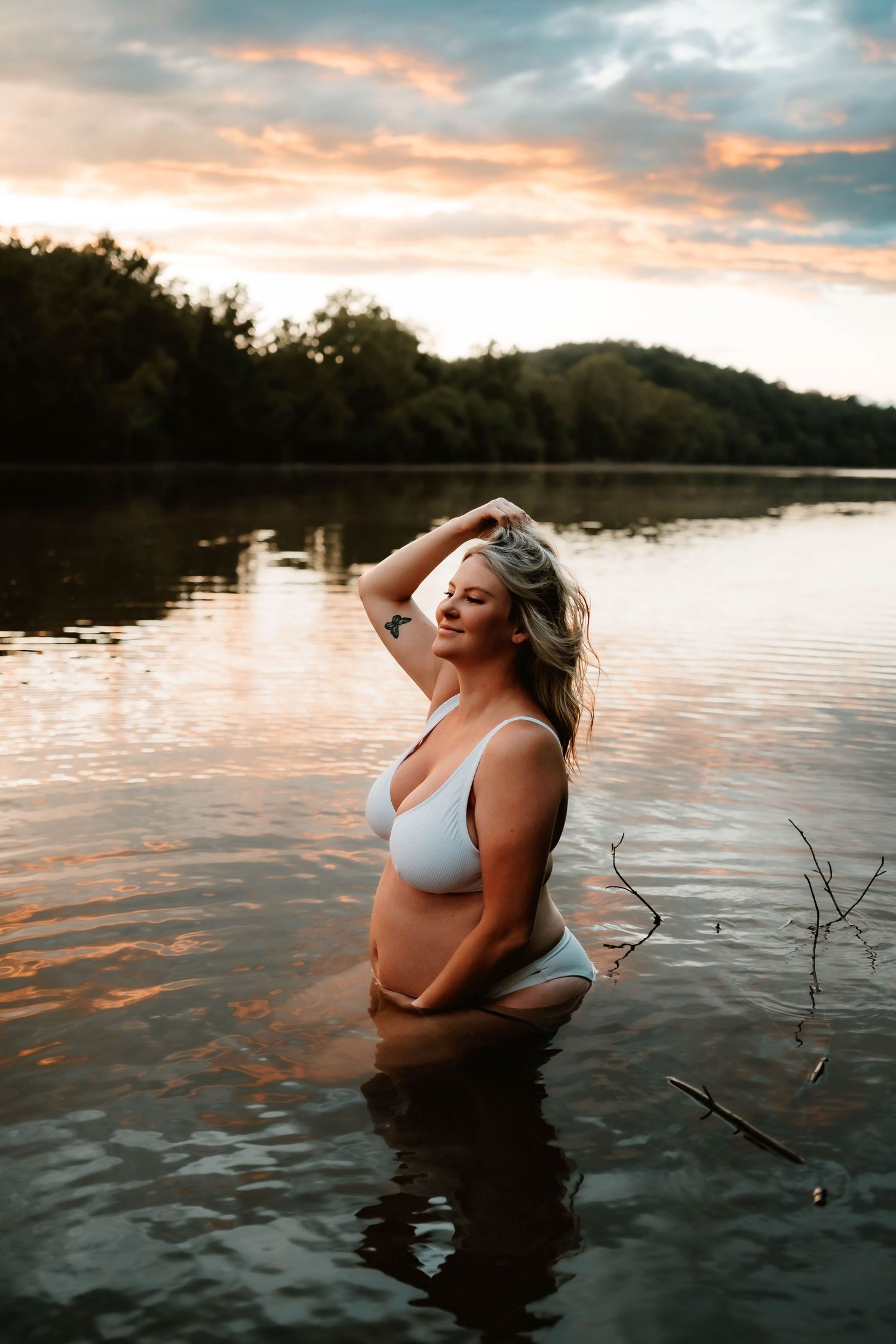 Woman in swimwear poses in lake water during golden sunset with mountain backdrop.
