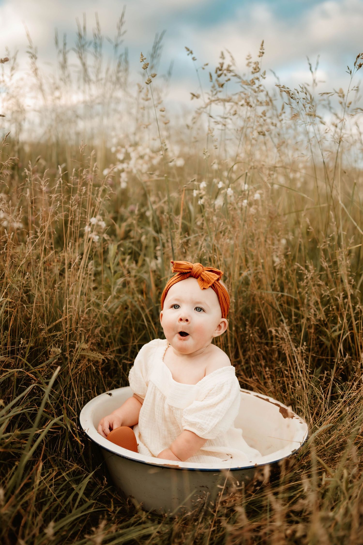 A baby in white dress and orange bow sits in vintage bathtub surrounded by tall grass.