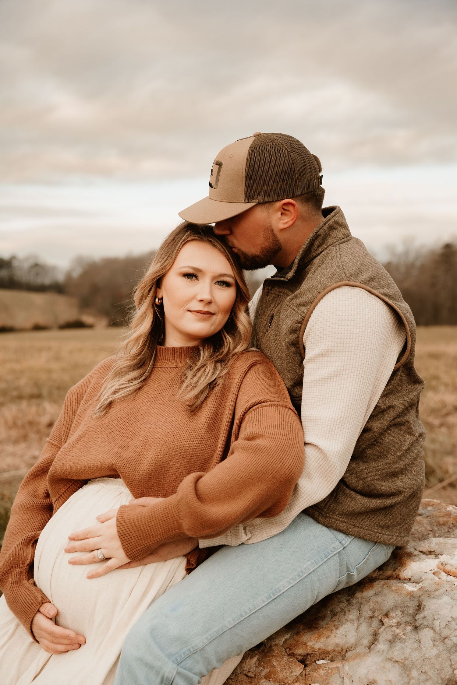 Romantic couple in autumn colors embraces on a rustic rock formation in a rural field at sunset.