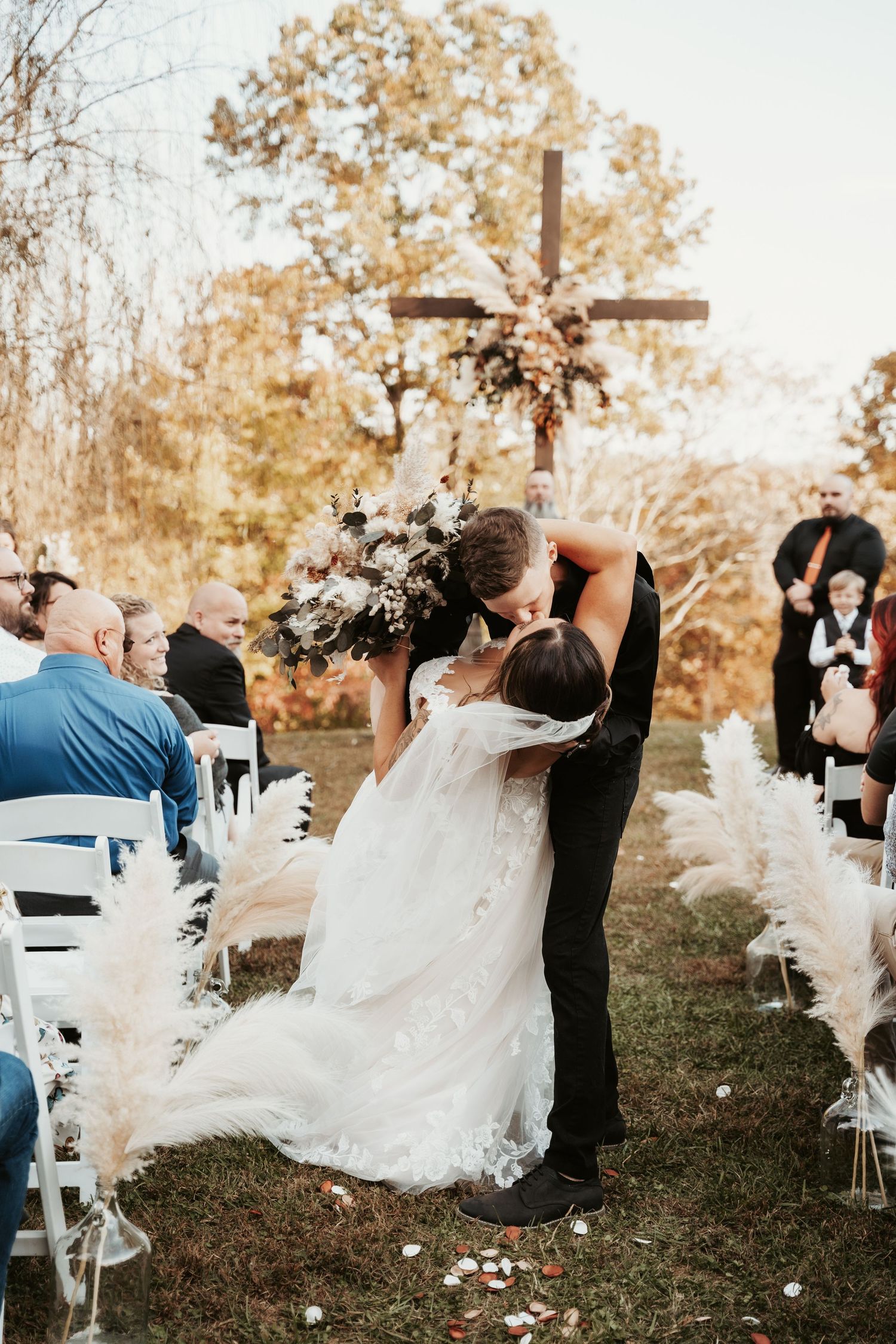 Wedding ceremony kiss at rustic outdoor venue with pampas grass aisle decor.