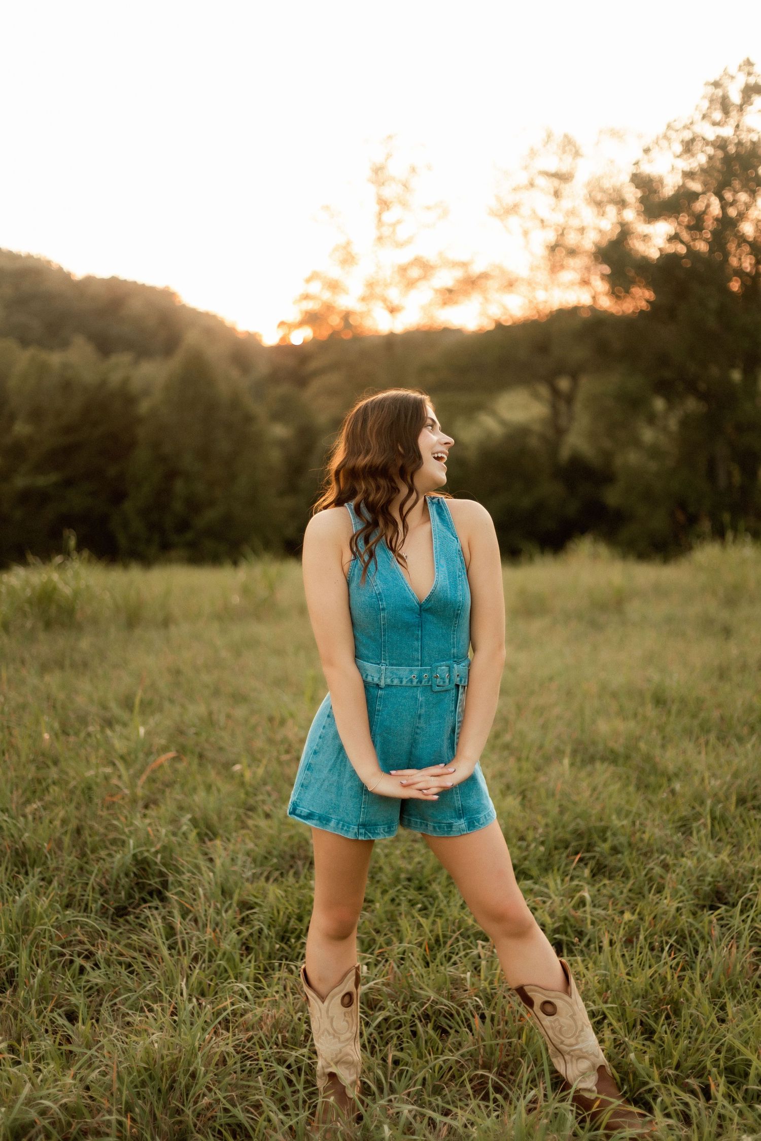 A young woman in a teal denim romper and cowboy boots laughs in a sunlit meadow at golden hour.