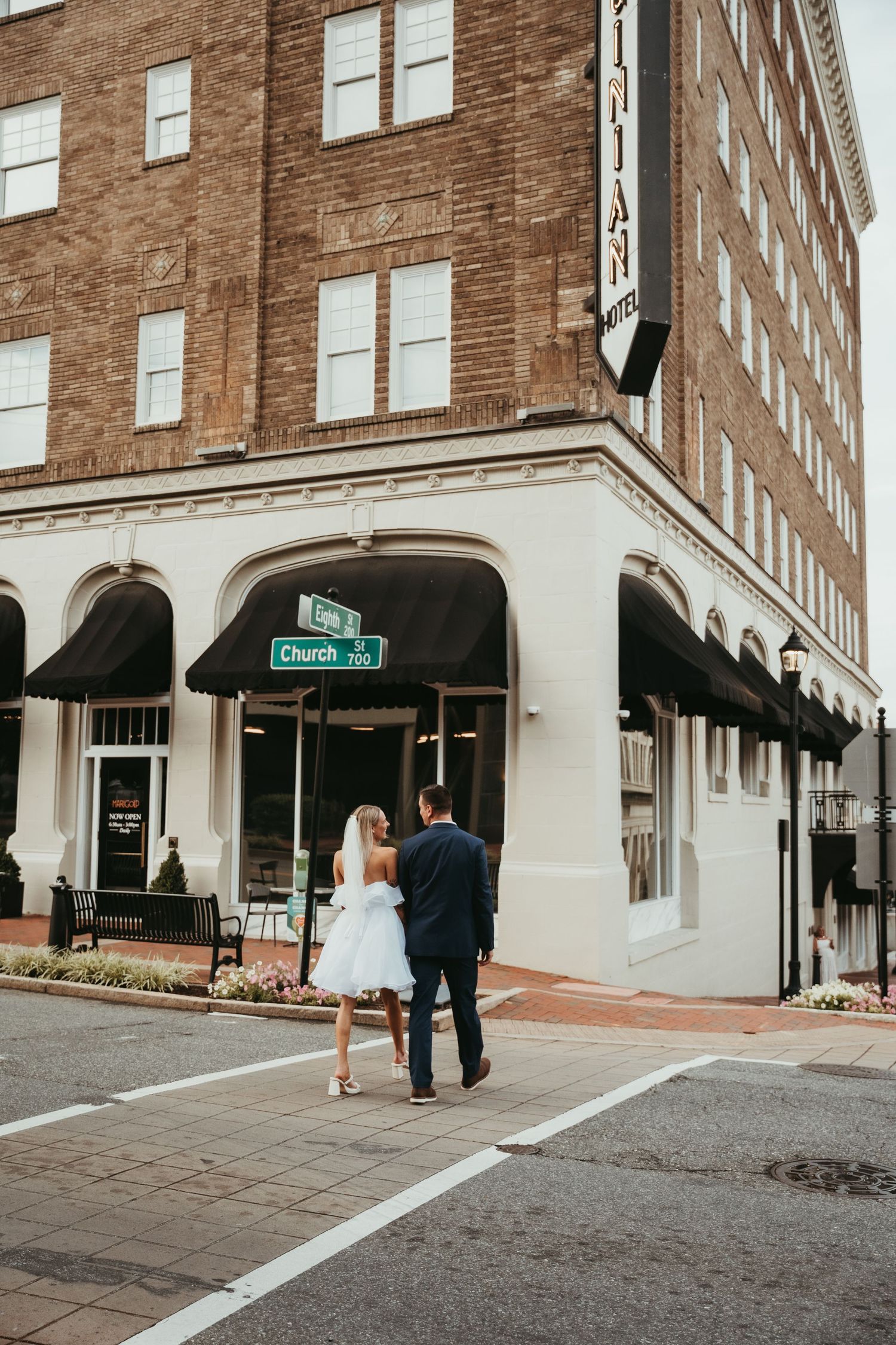 Wedding couple exits historic brick building with classic architecture and black awnings.