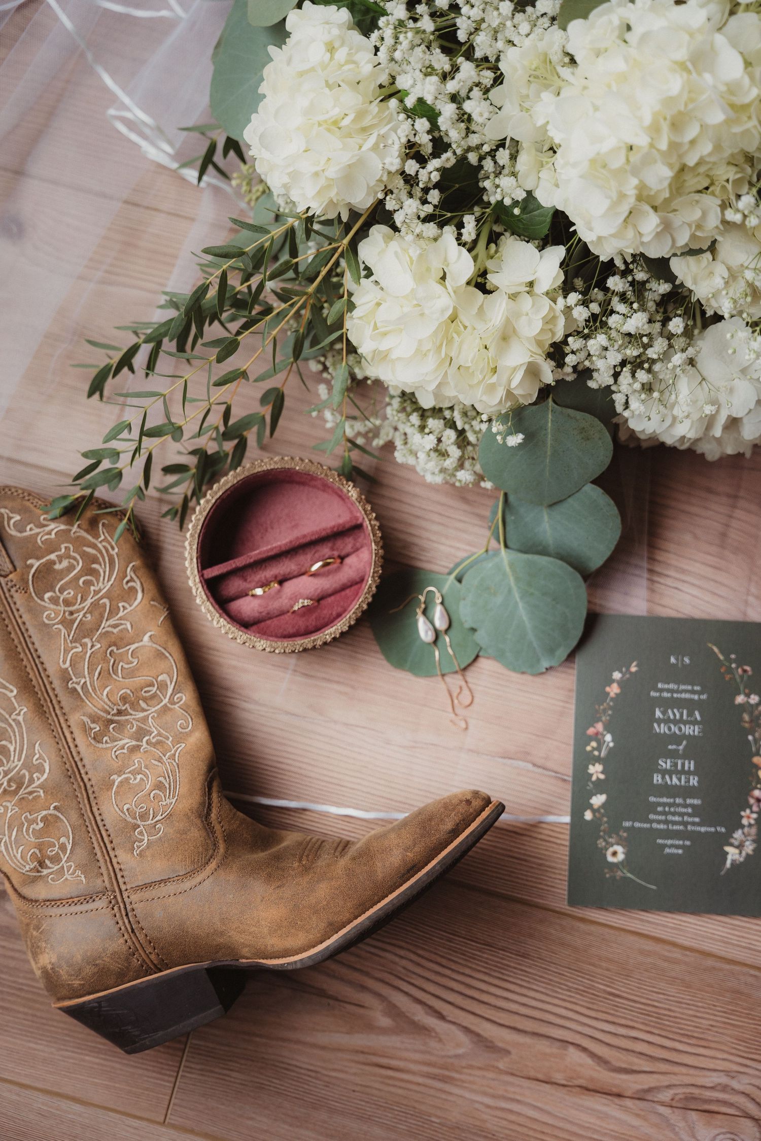 Wedding details featuring brown cowboy boots, white flowers, and invitation cards on wooden surface.