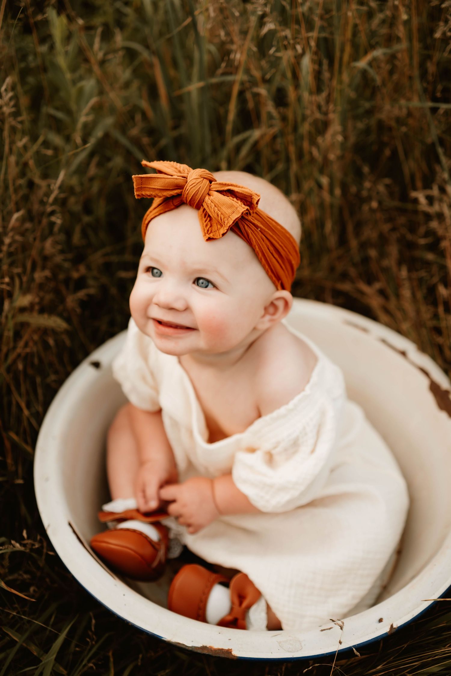 A smiling baby in white dress and orange headband sits in antique washbasin outdoors.