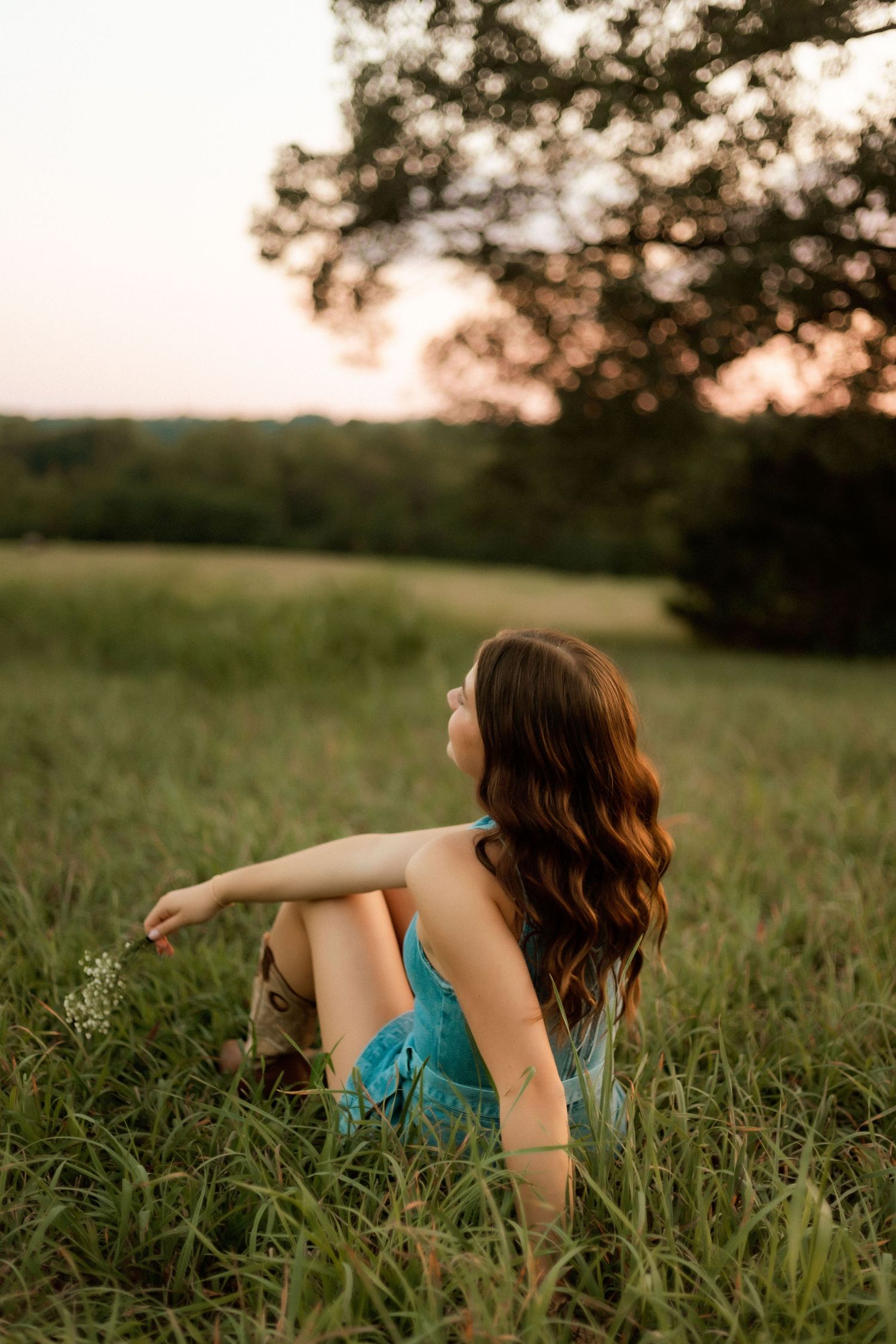Young woman in teal dress sitting in green meadow at sunset, with long wavy hair, gazing toward golden sky.