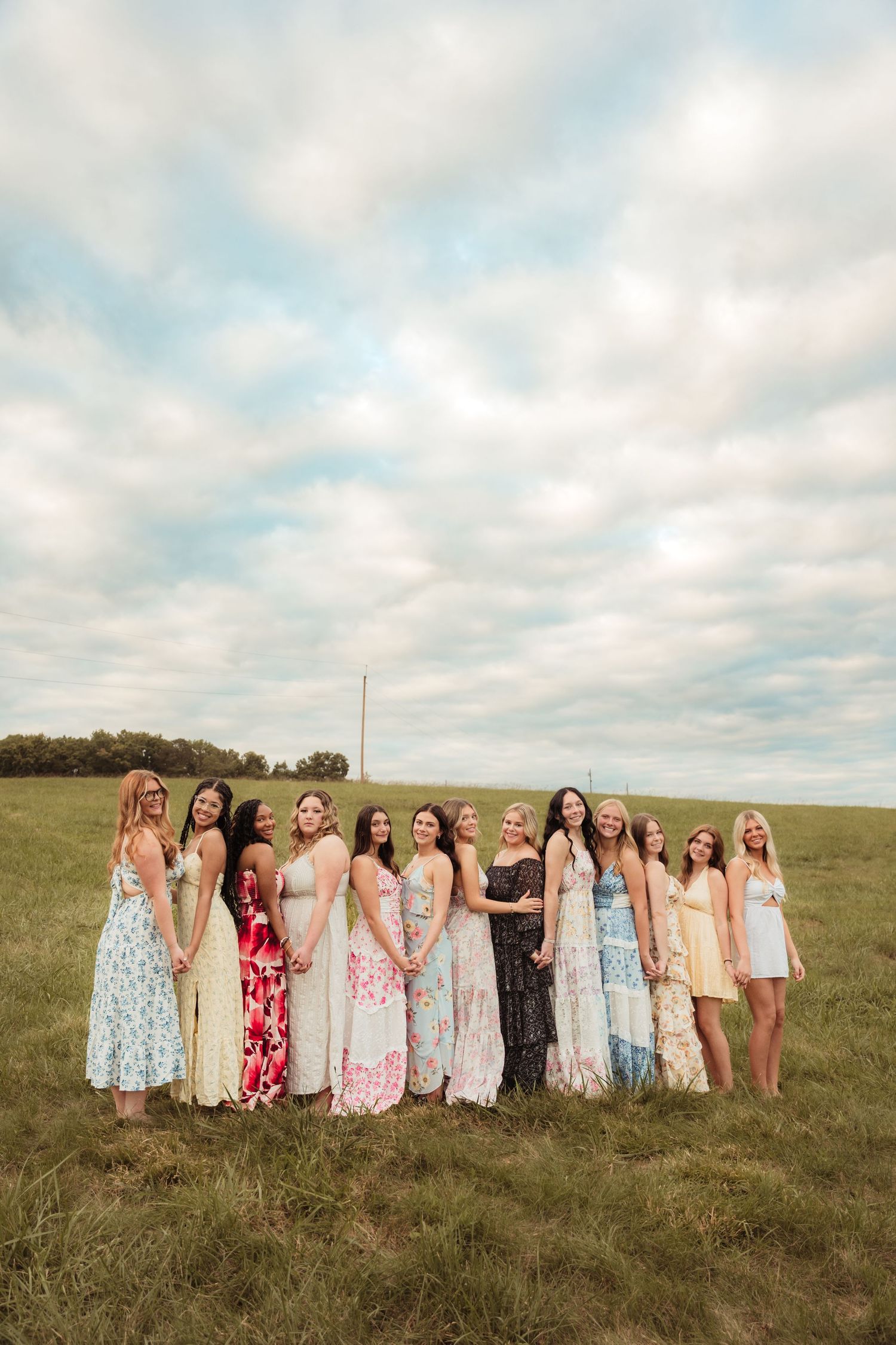 Group of women in colorful floral maxi dresses posing together in a green field under a cloudy sky.