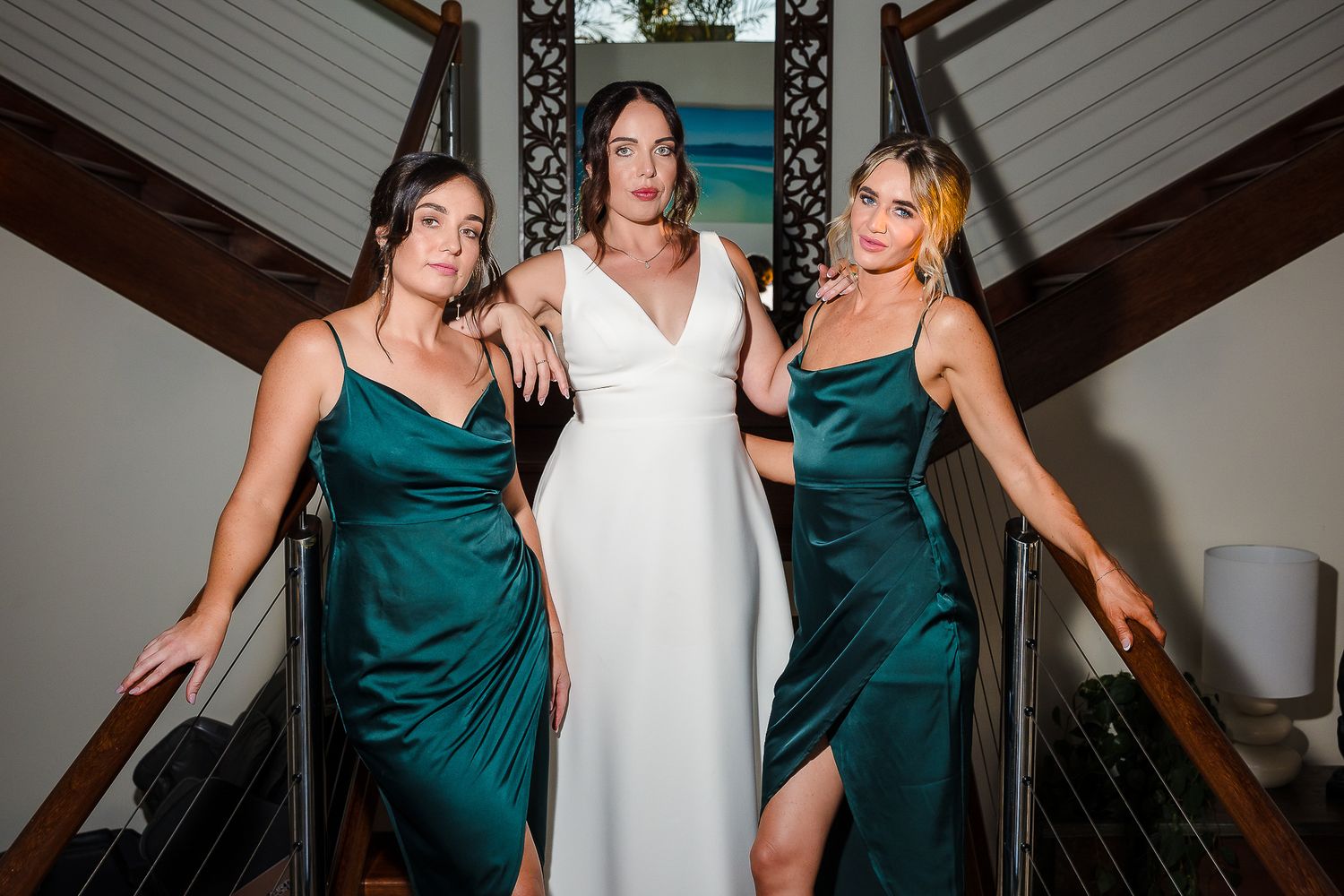 Three women pose on staircase wearing elegant emerald green and white formal dresses.
