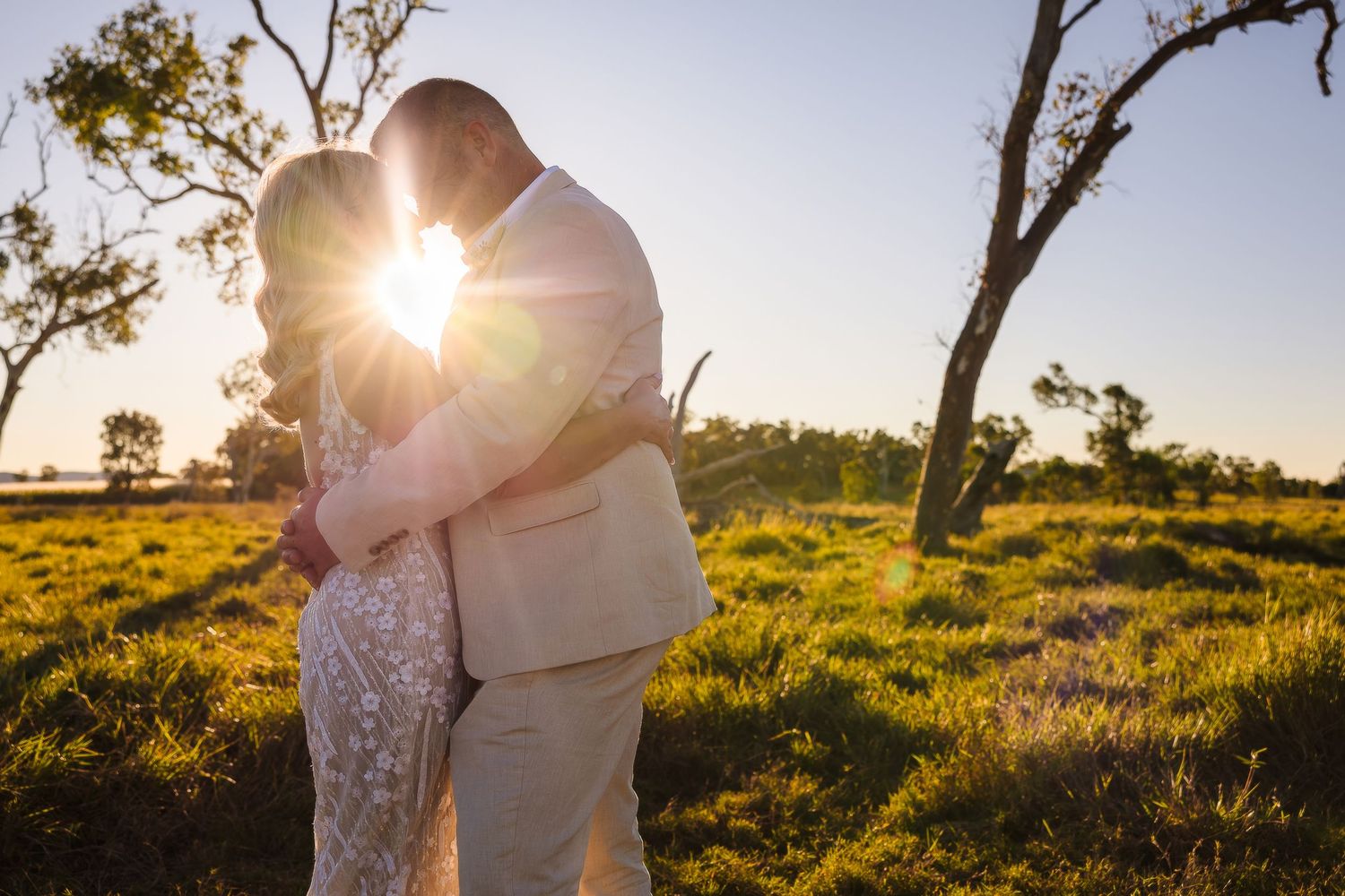 A couple in formal attire embrace while backlit by golden sunlight in a grassy field at sunset.