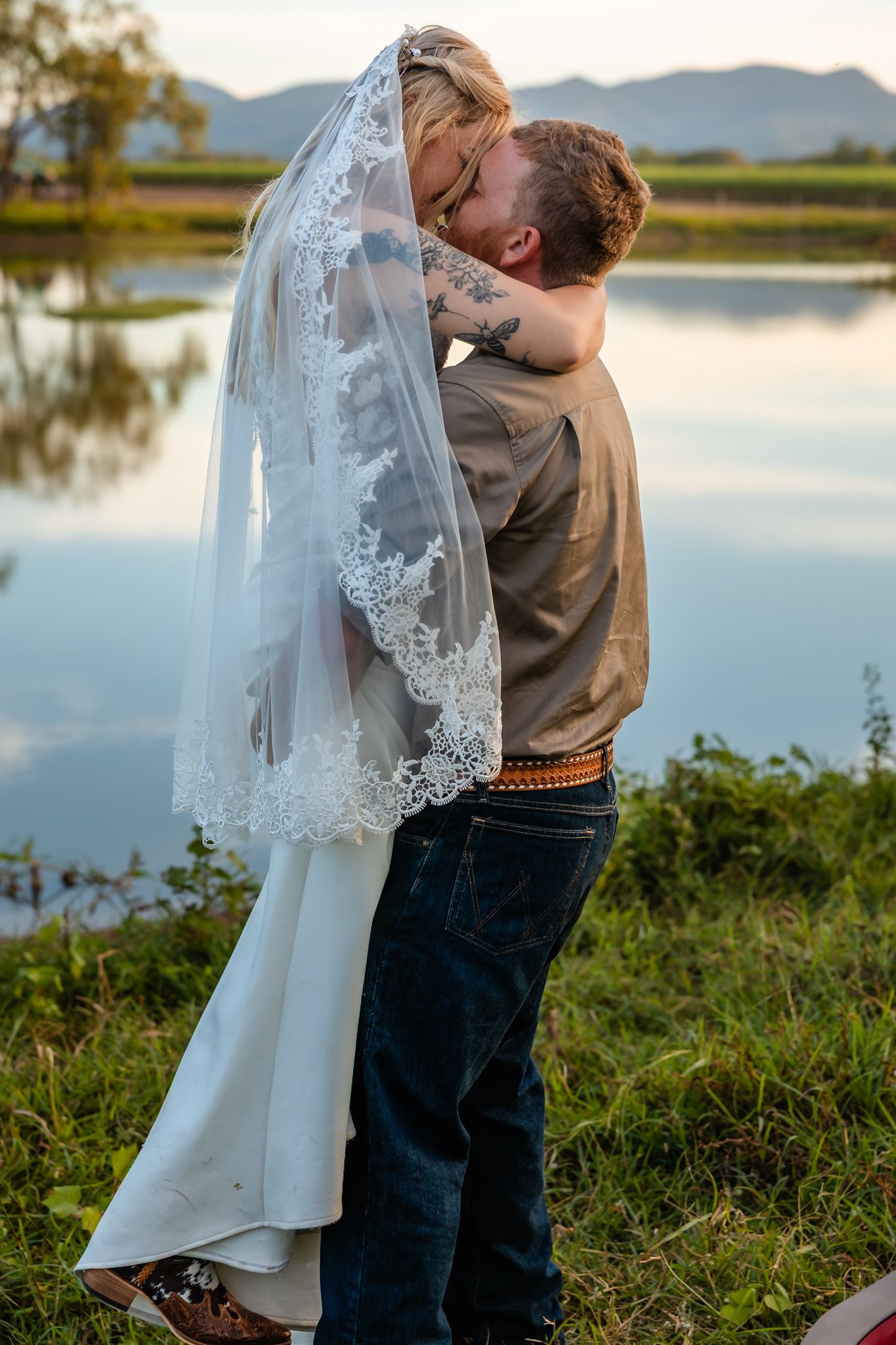 Bride and groom sharing a romantic kiss under a lace veil by a reflective lake at sunset.