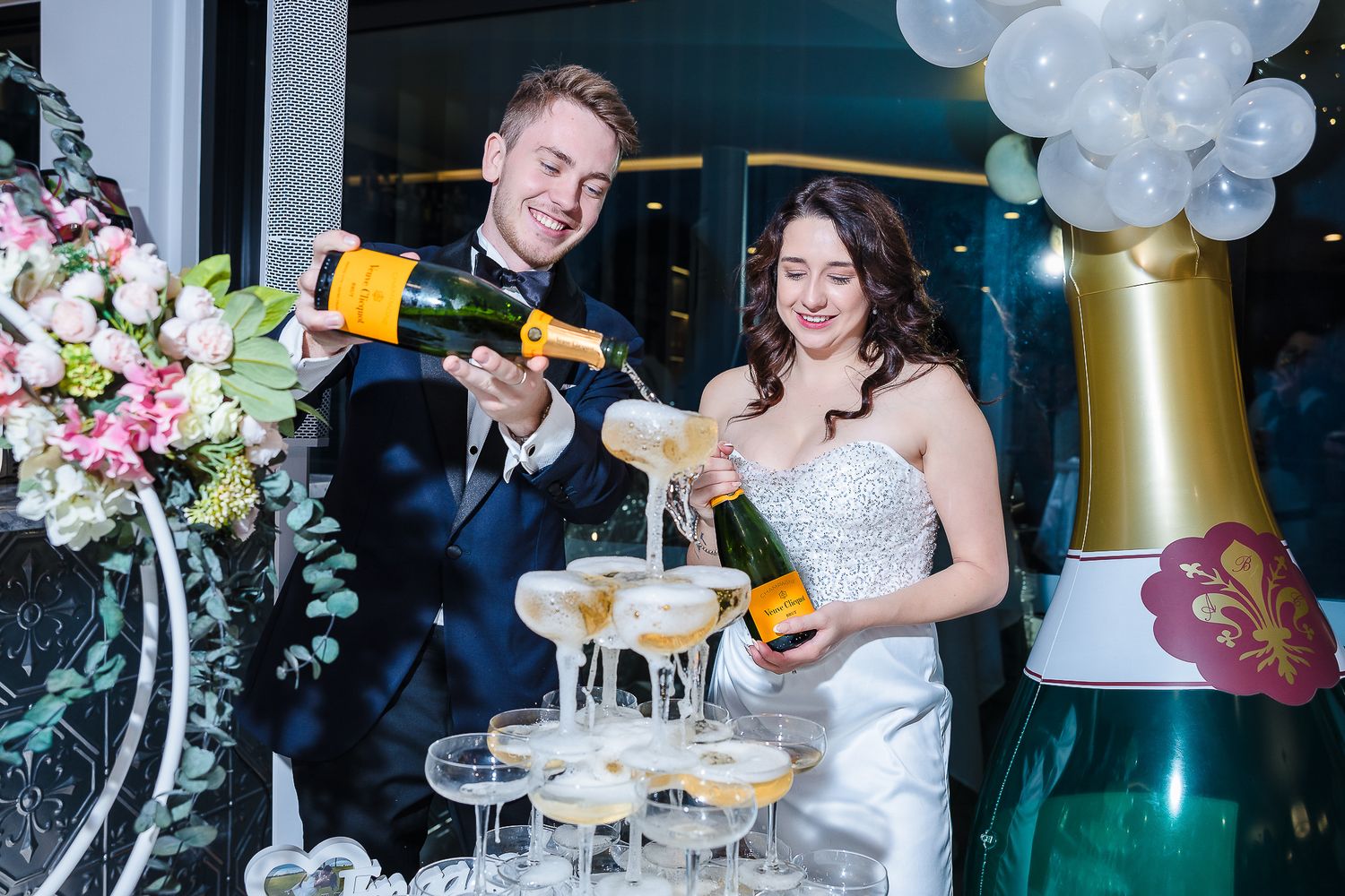 Couple pours champagne into tower of glasses at wedding reception with balloon decor.