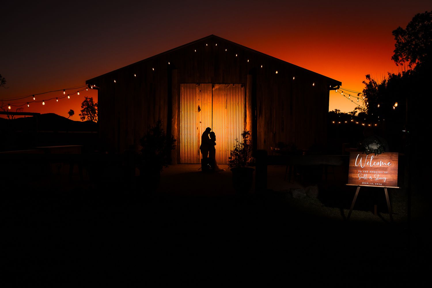 Silhouette figure stands in front of a barn with string lights against a dramatic orange sunset sky.