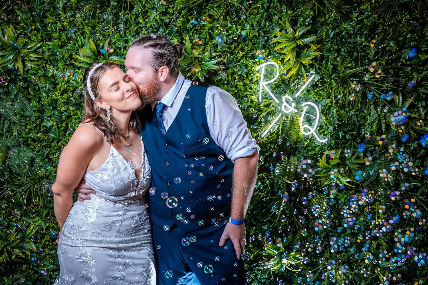 Couple poses against green hedge wall with neon sign and floating bubbles.
