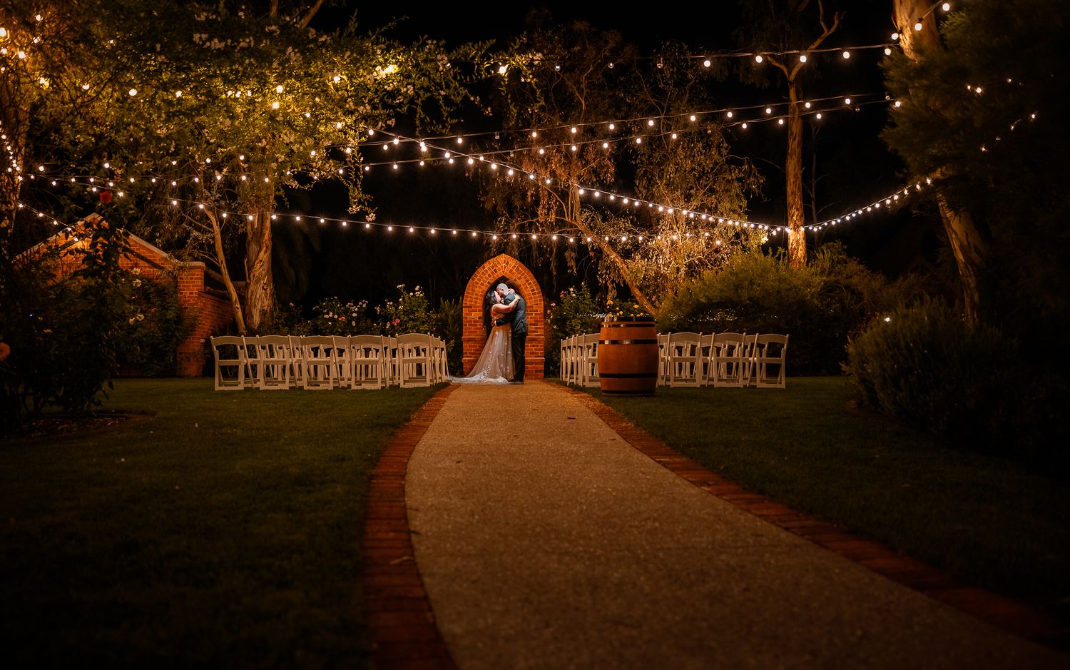 Romantic pathway lit by string lights leads to rustic barn wedding venue at night.