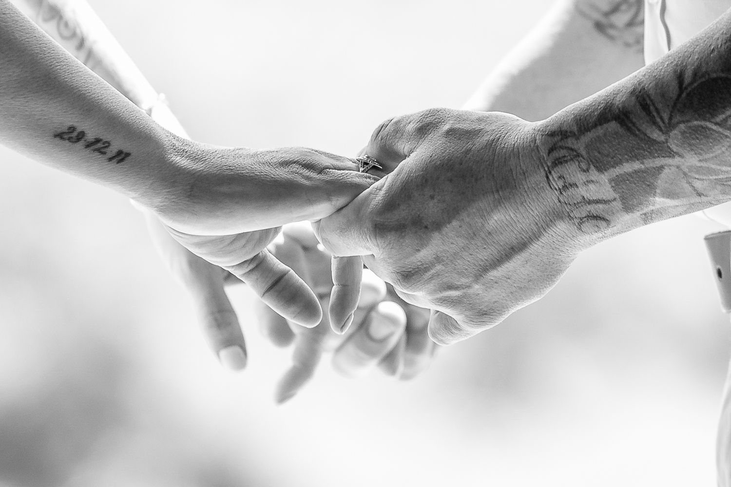 Black and white close-up of tattooed hands holding each other tenderly.