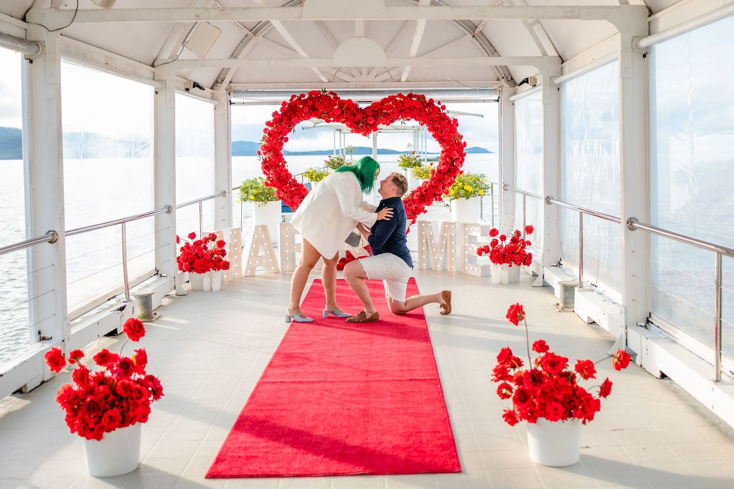 Romantic proposal scene on a boat deck with red heart balloon arch, red carpet, and floral arrangements.