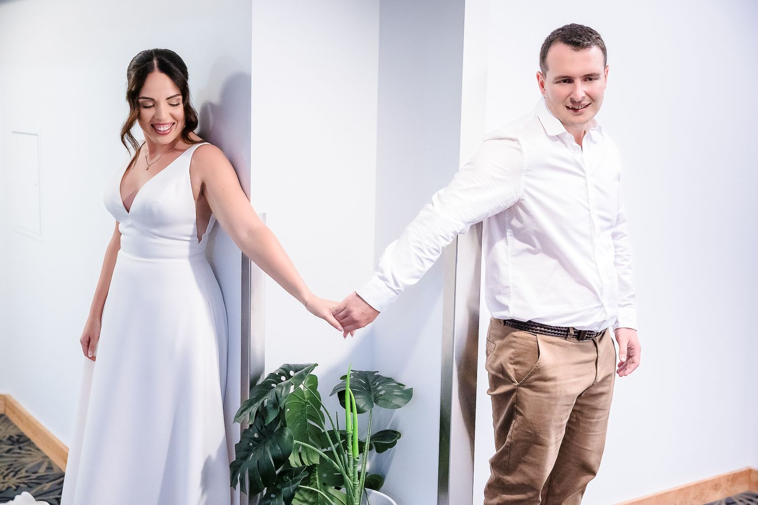 A couple stands separated by a white wall while playfully holding hands in a wedding photoshoot setting.