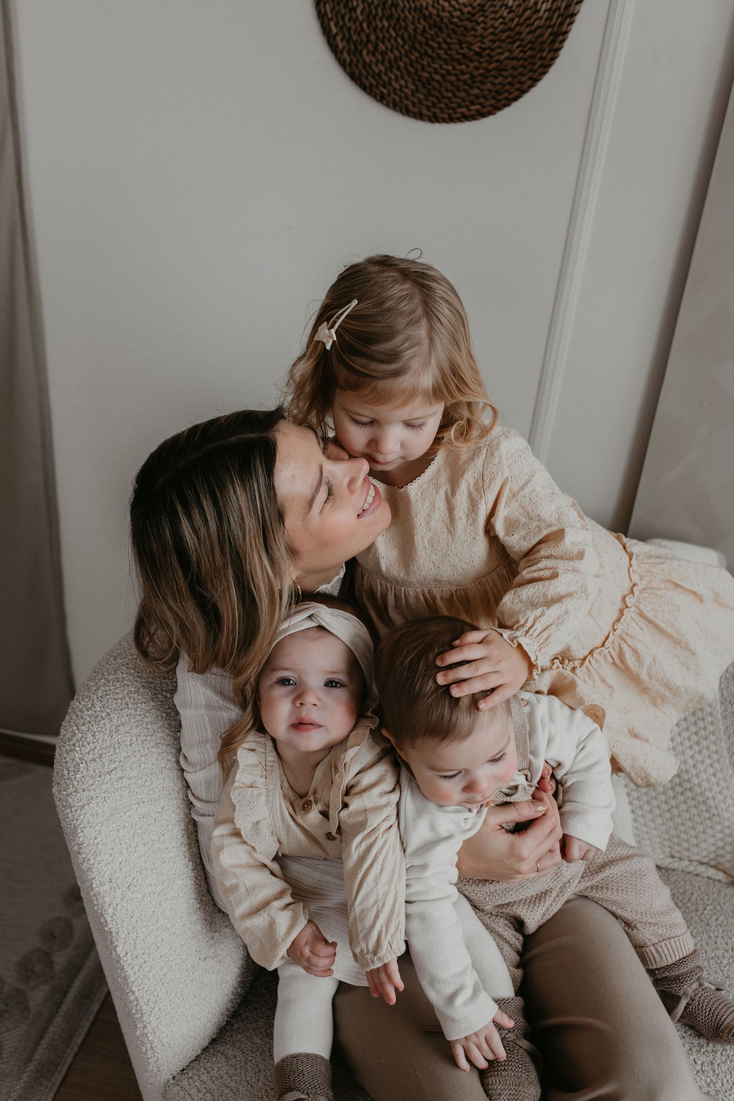 Glimlachende moeder in een fauteuil omringd door haar drie jonge  kinderen in lichte beige kleding tijdens een binnenfotoshoot.