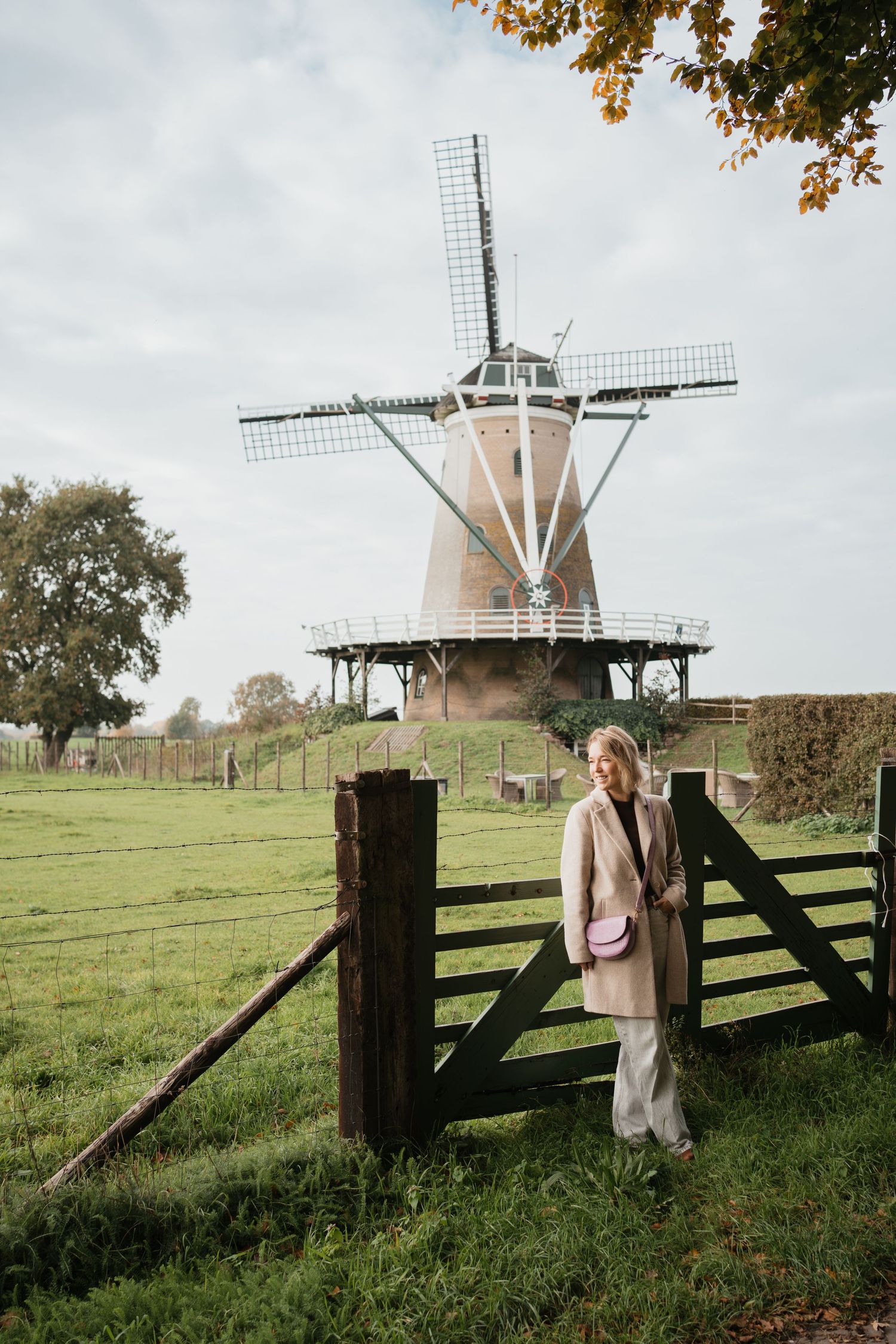 Blonde vrouw in beige jas met roze tas bij het hek van molen De Windhond in Soest, met een groen weiland op de voorgrond.