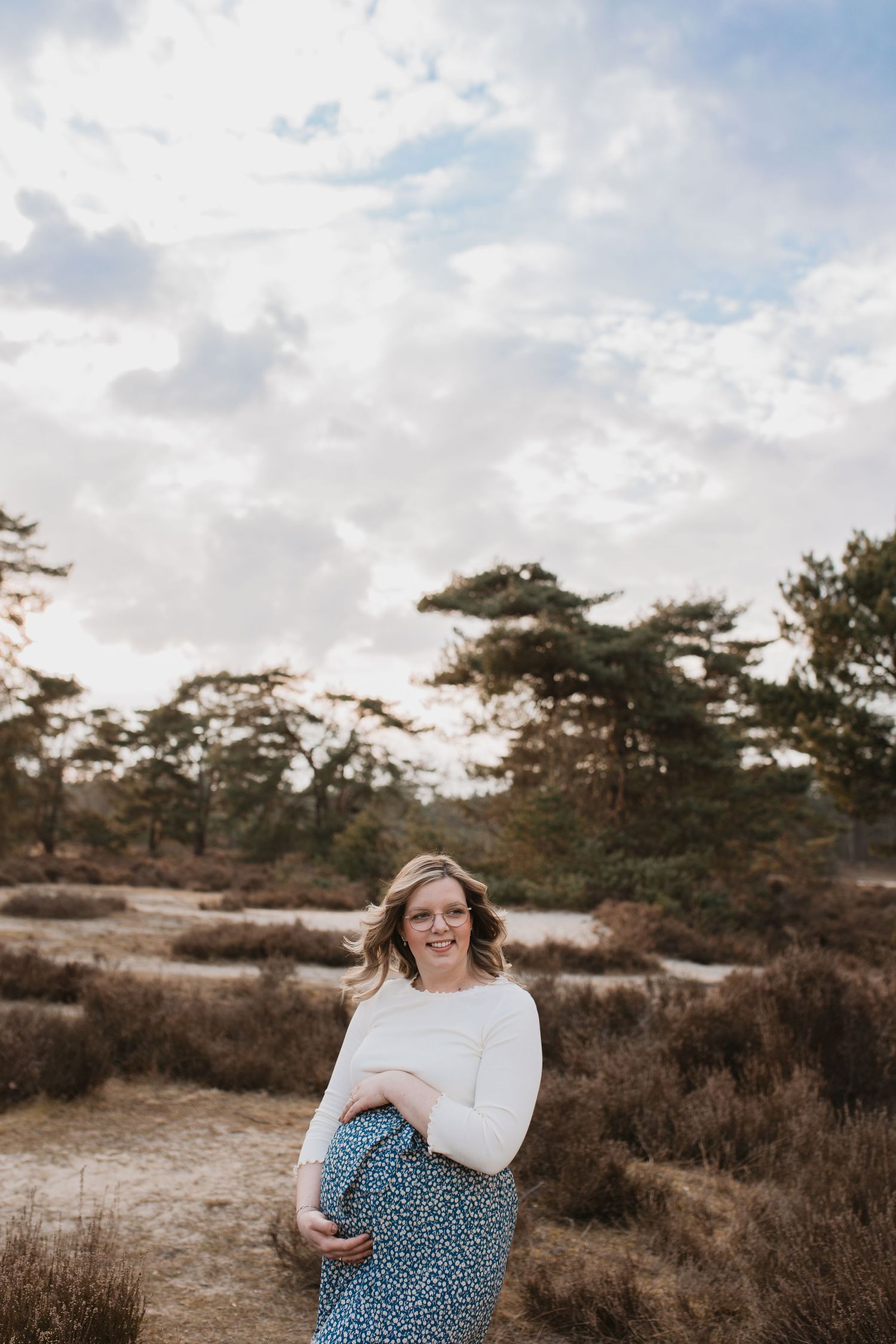 Zwangere vrouw in een witte top en blauw geprinte rok glimlacht en houdt haar buik vast tijdens een shoot tussen de heide in Soest.
