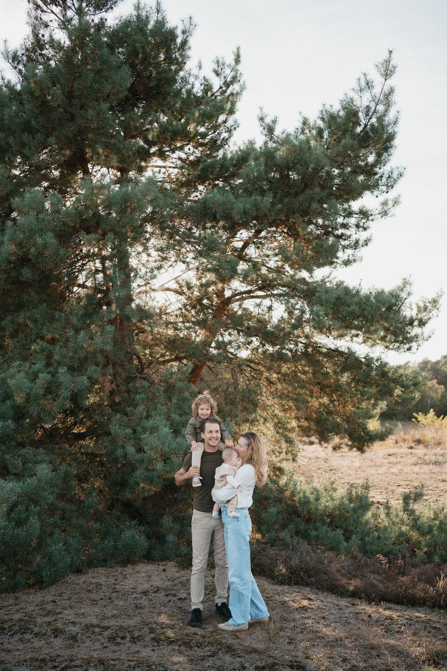 Gezin samen in de natuur bij een grote dennenboom, met grote zus op de schouders van vader en de jongste dochter in de armen van mama.