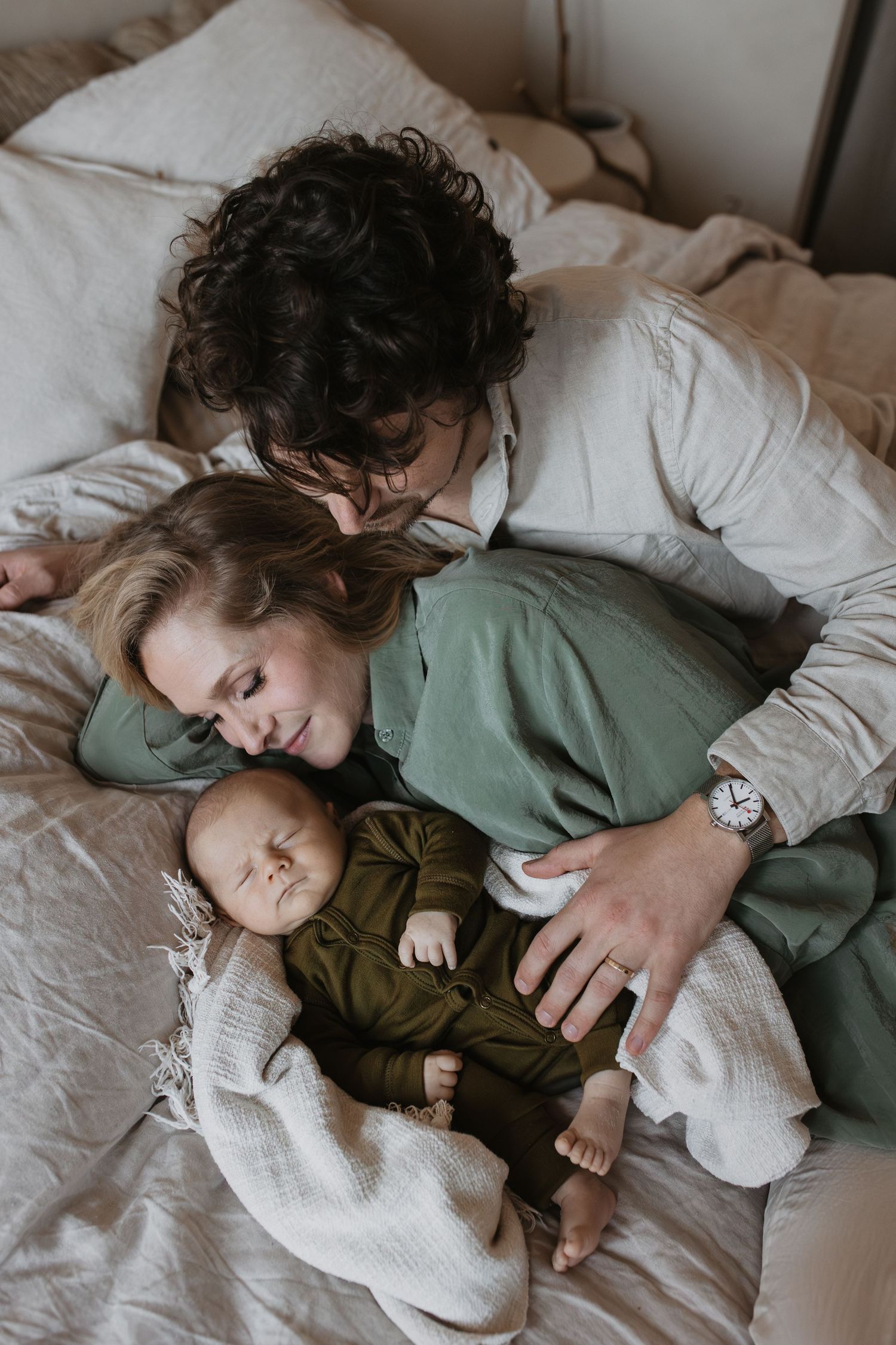 Newbornshoot op bed. Een knuffelende mama en papa liggen dicht tegen elkaar aan geknuffeld achter hun slapende baby.