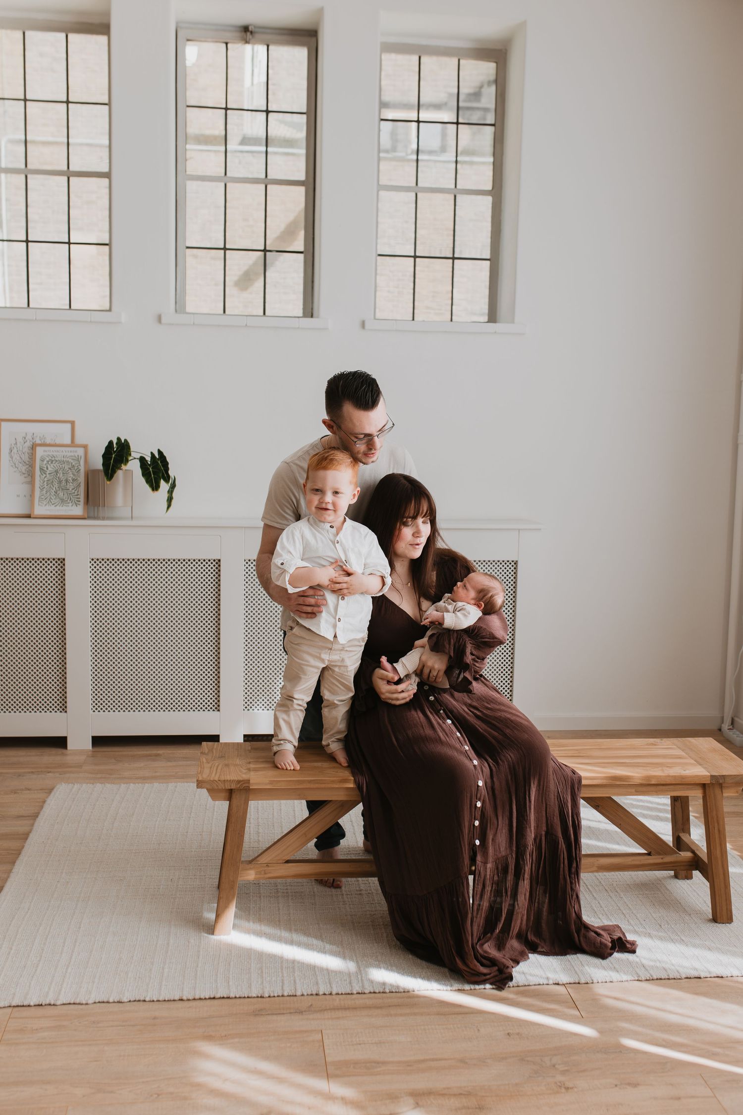 Gezinsfoto in een lichte studio, vader houdt de peuter vast, zittende moeder in een bruine jurk hout de newborn baby vast.