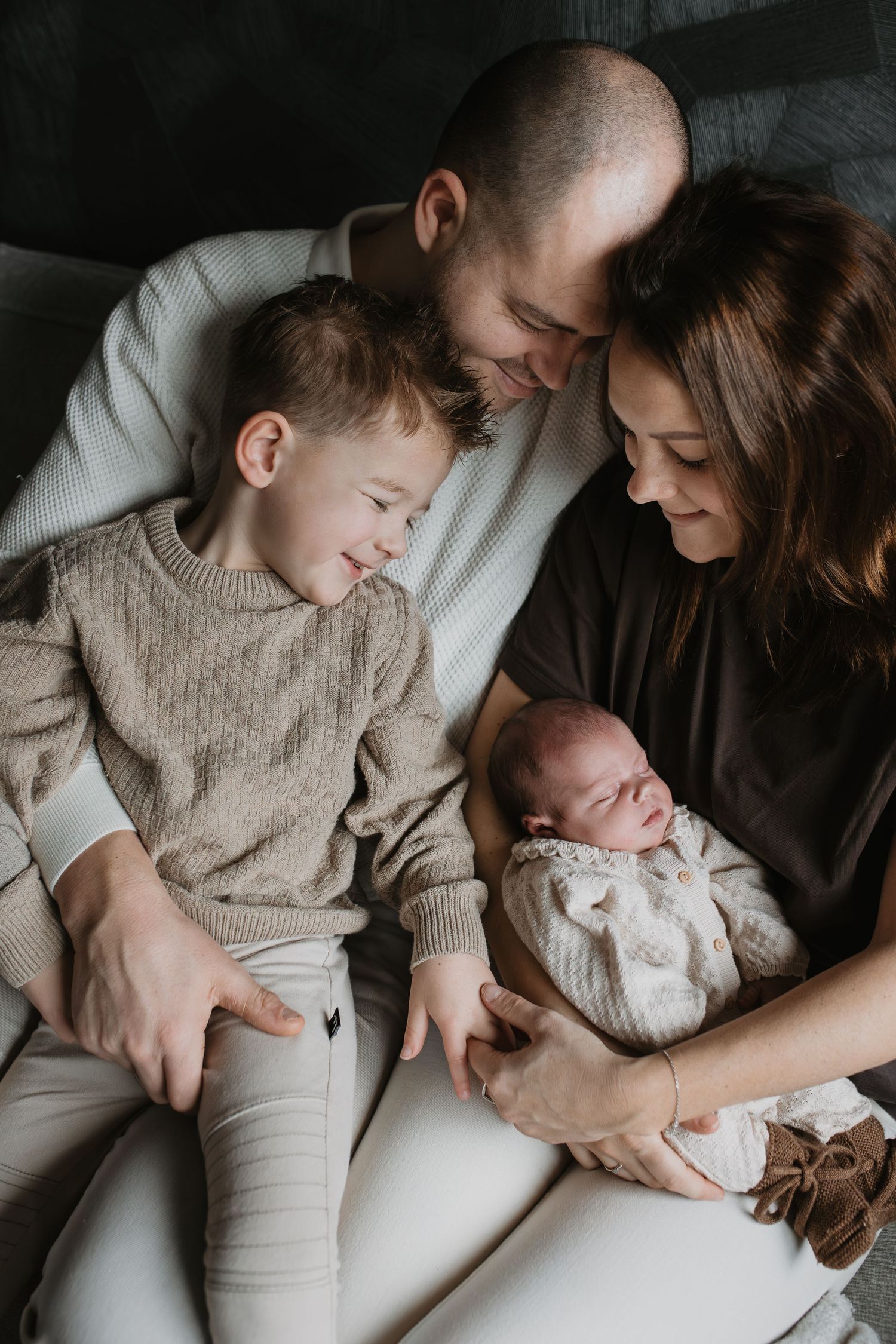 Papa en mama houden hun hoofden tegen elkaar, terwijl hun zoon en de baby knus in het bruin voor hen zitten.