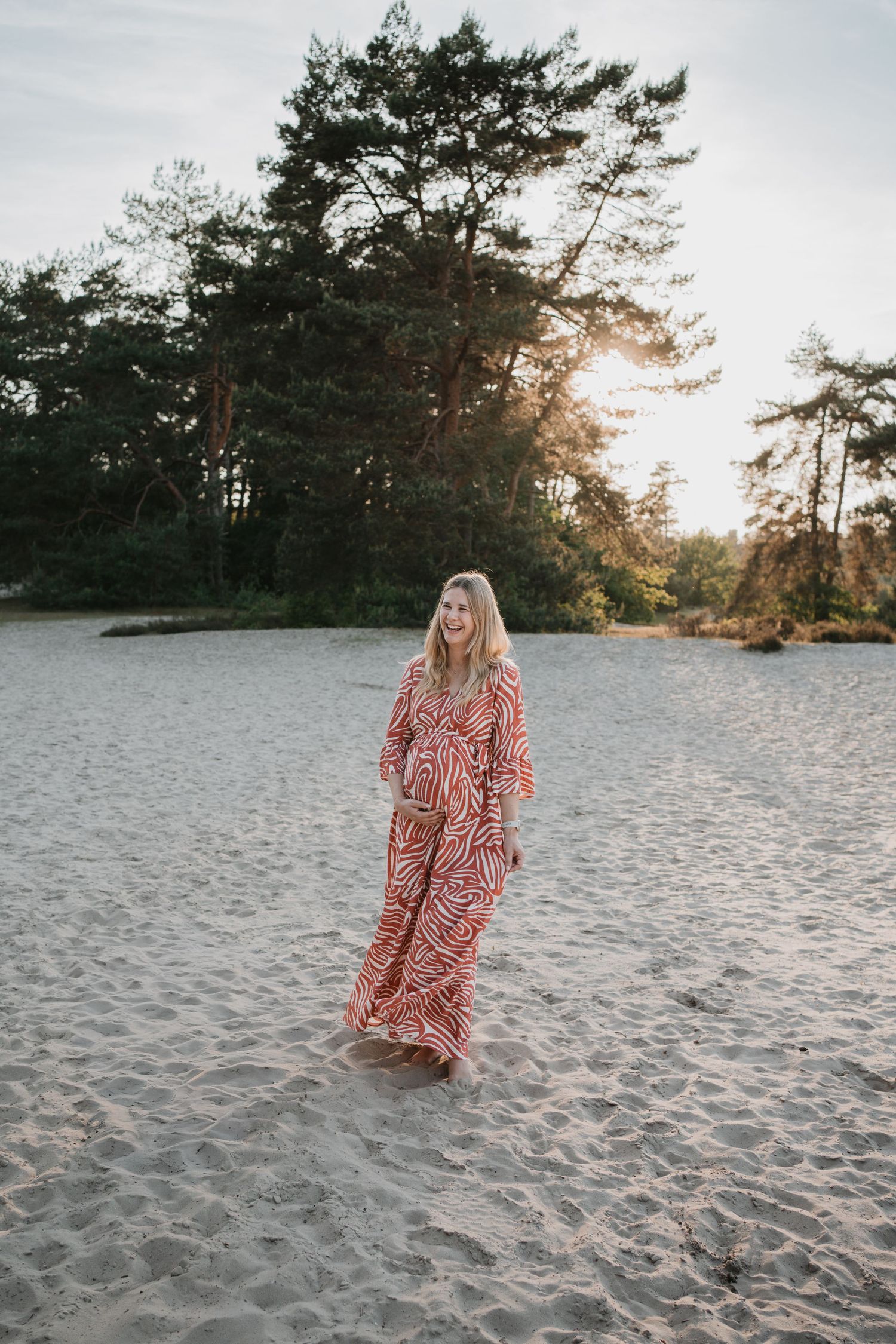 Blije zwangere vrouw in een lange rode jurk lacht tijdens een zwangerschapsshoot op een zandvlakte in de natuur.