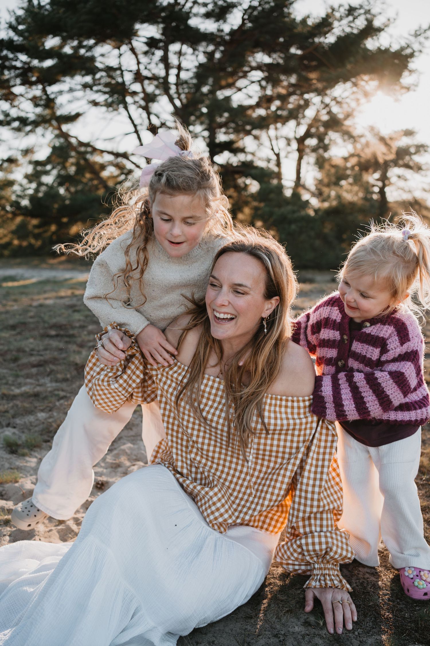 Mama lacht en haar twee dochters hangen liefdevol om haar nek op de Soester Duinen tijdens haar gezinsshoot.