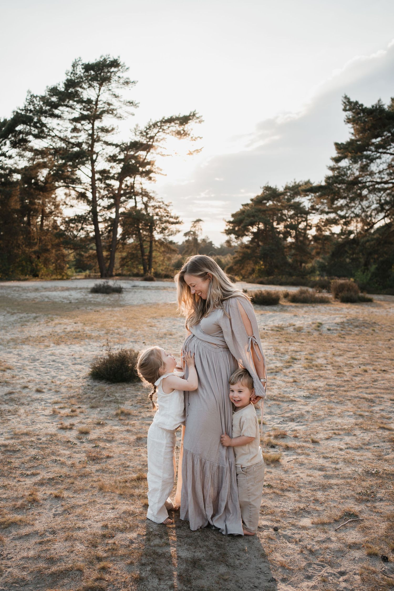 Een zwangere vrouw in een lange grijze jurk staat glimlachend met twee jonge kinderen in een natuurgebied met bomen tijdens zonsondergang.
