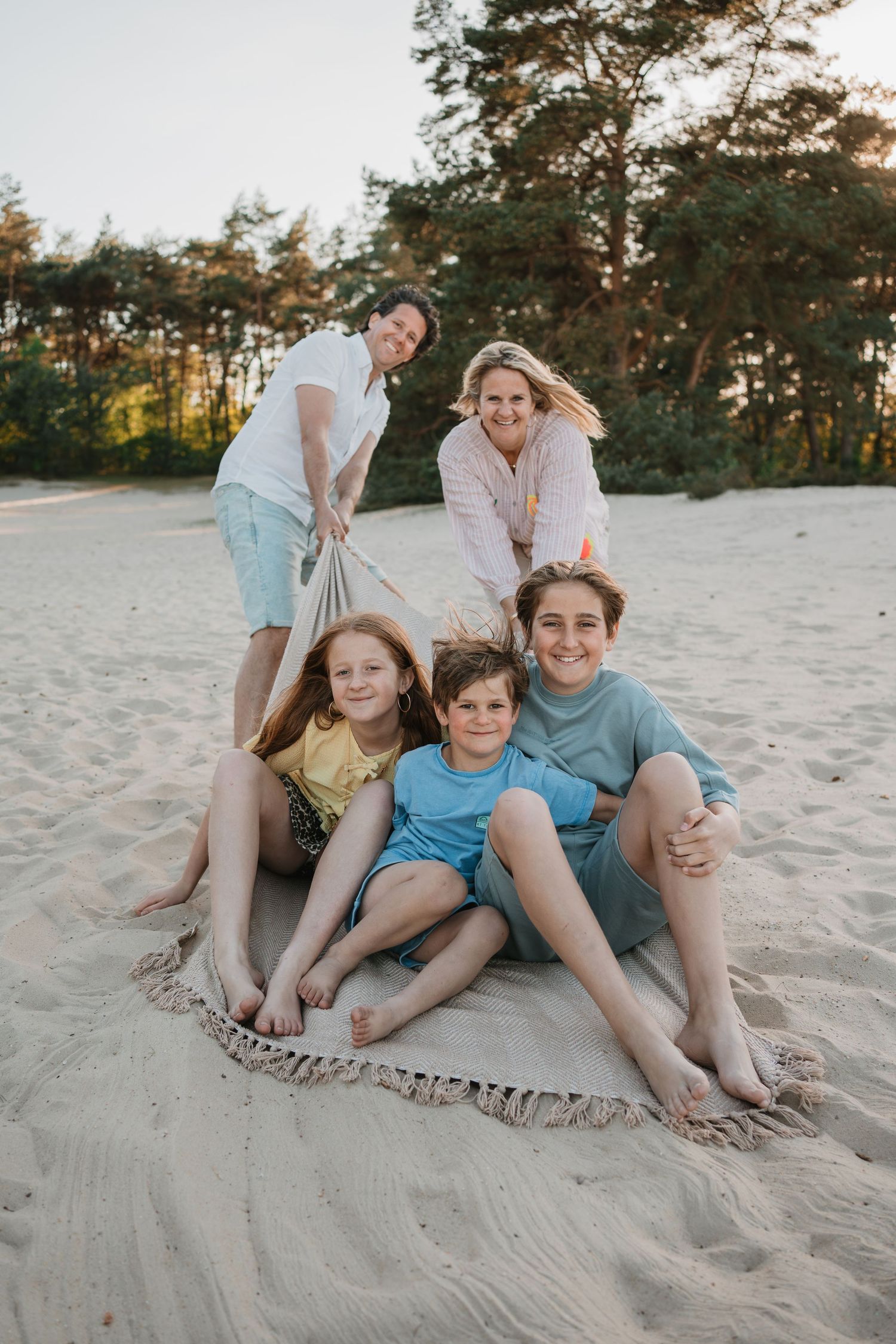 Lachende ouders trekken hun drie kinderen speels vooruit op een picknickkleed in een zandverstuiving bij het bos.