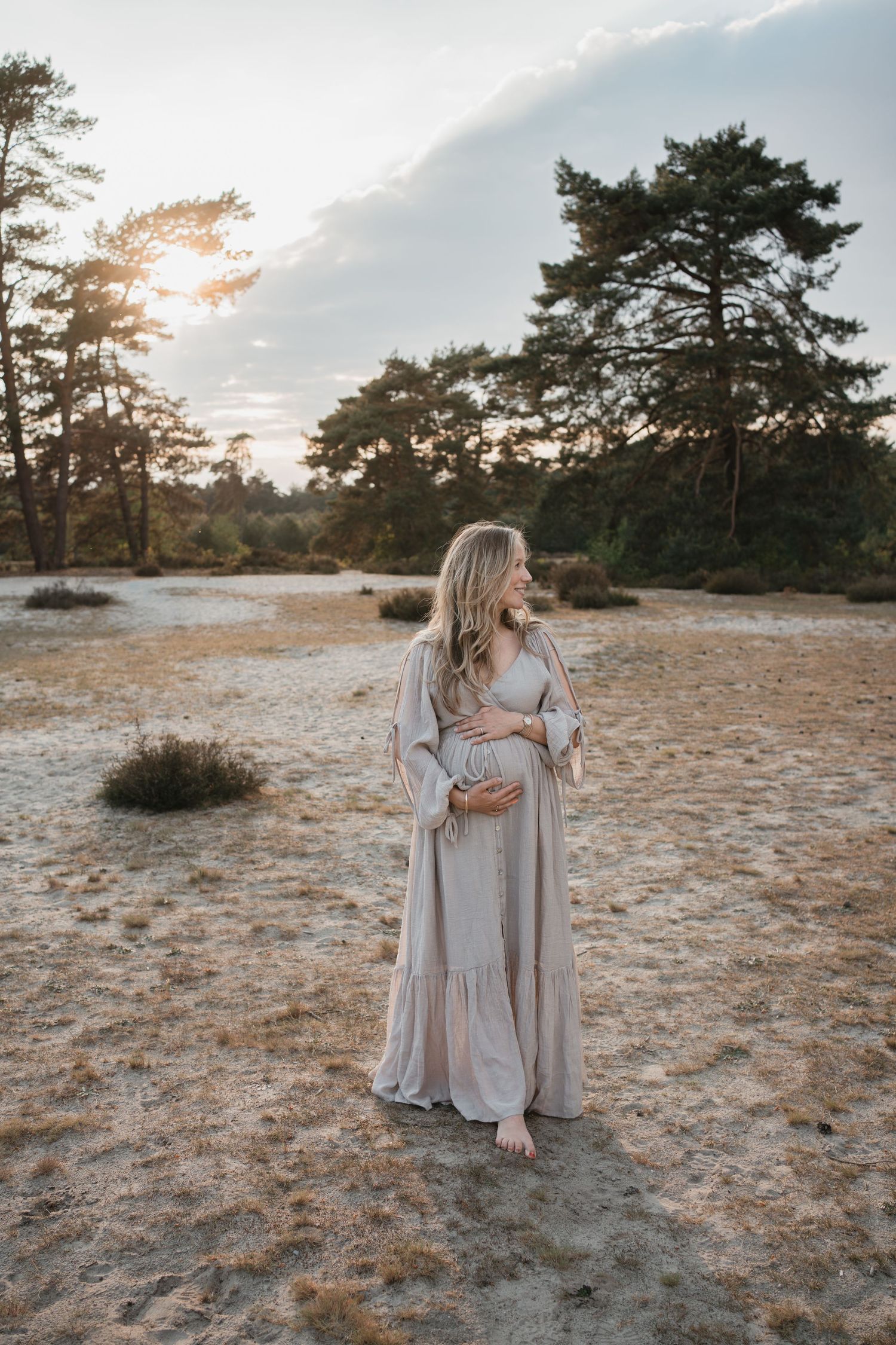 Een zwangere vrouw in een lange grijze jurk houdt haar buik vast op een zandvlakte in Soest tijdens een fotoshoot bij golden hour.