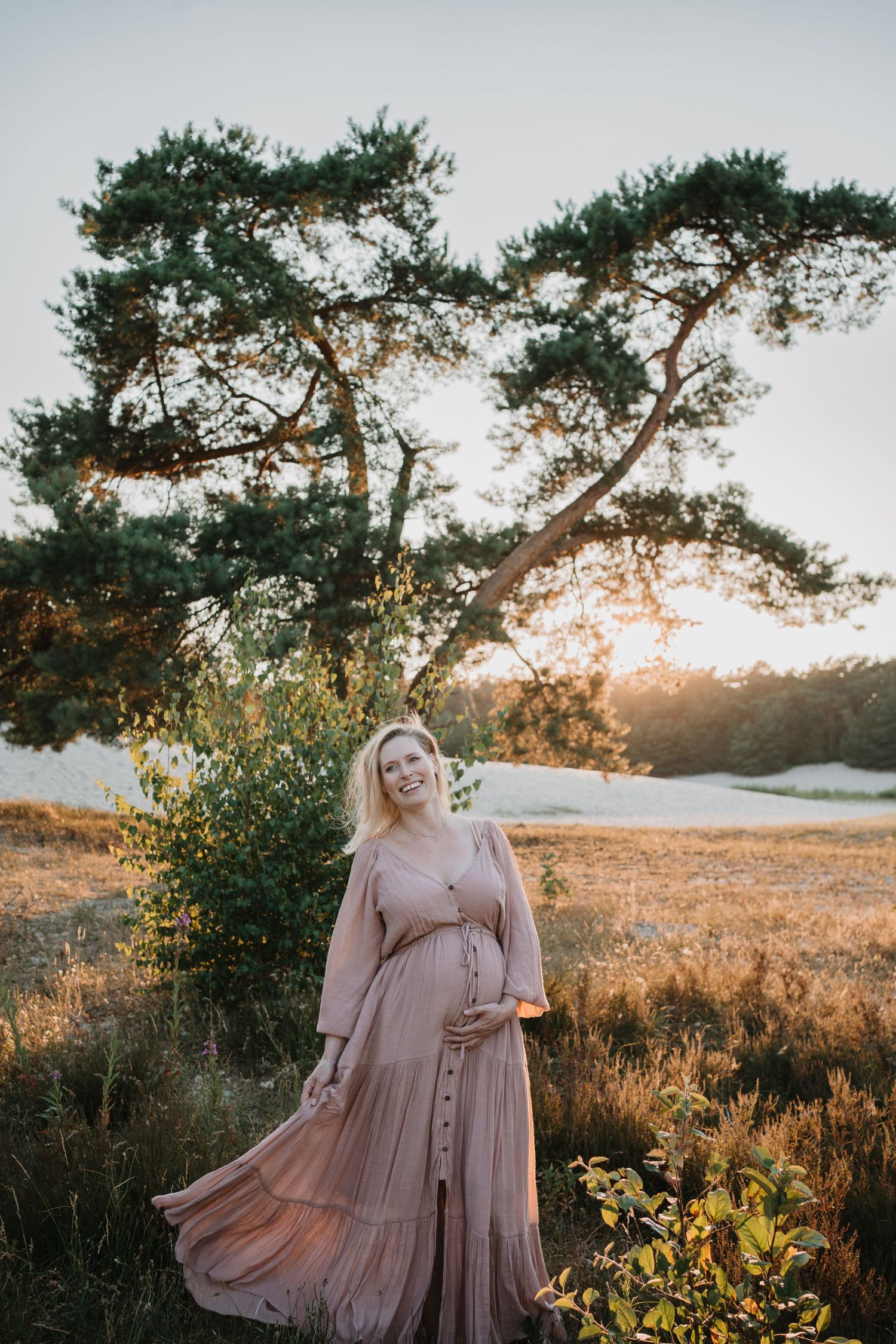 Lachende zwangere vrouw in een roze jurk op een heideterrein bij zonsondergang, gefotografeerd tijdens een zwangerschapsfotoshoot.