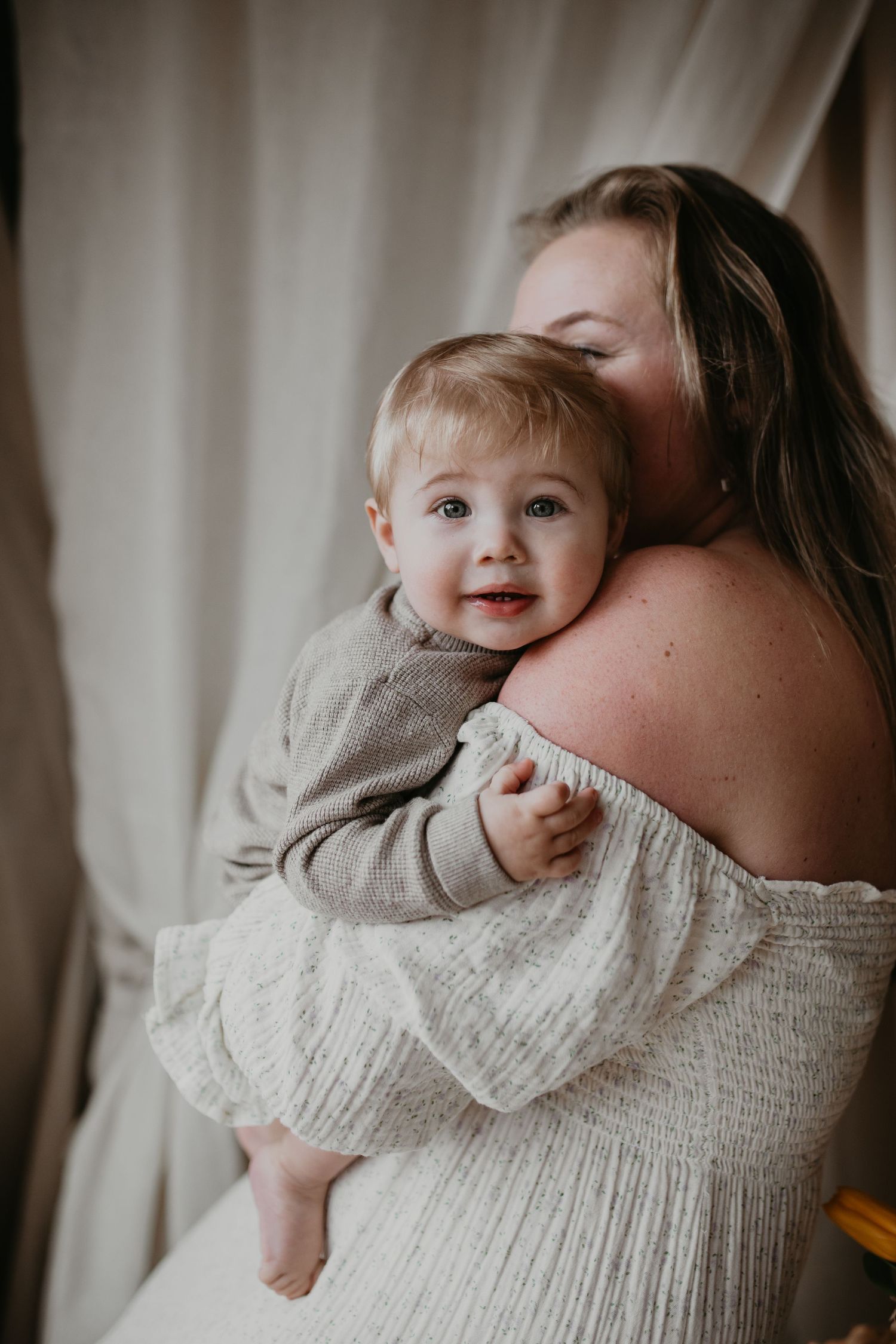 Glimlachende vrouw houdt jongetje met heldere blauwe ogen en blond haar vast in haar armen. Jongetje kijkt recht in de camera