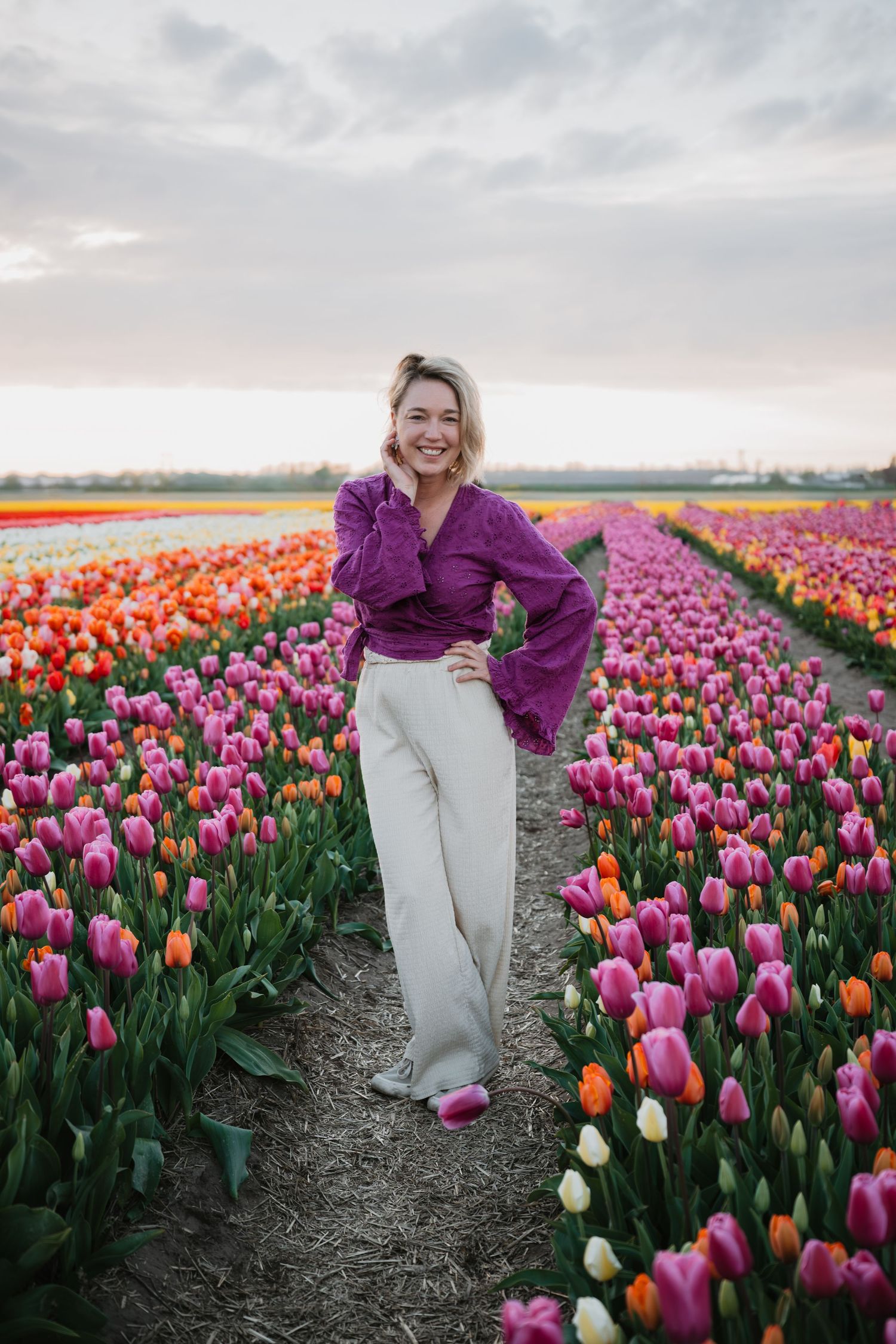 Glimlachende vrouw in een paarse top en beige broek staat in een kleurrijk tulpenveld onder een bewolkte lucht.