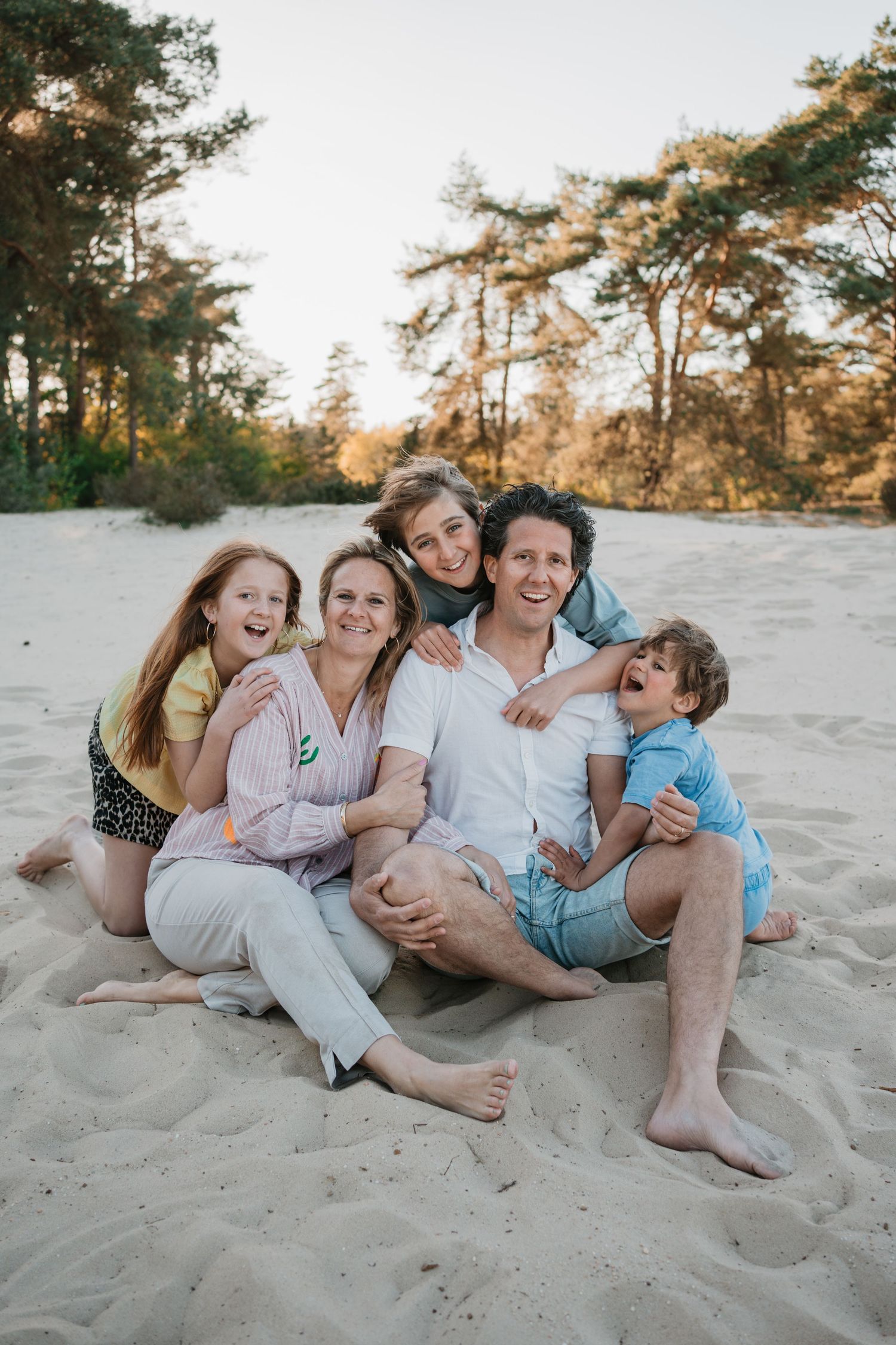Gezinsfotograaf, een vrolijk gezin met drie kinderen zit lachend bij elkaar in het zand van de Soester Duinen tijdens een fotoshoot.