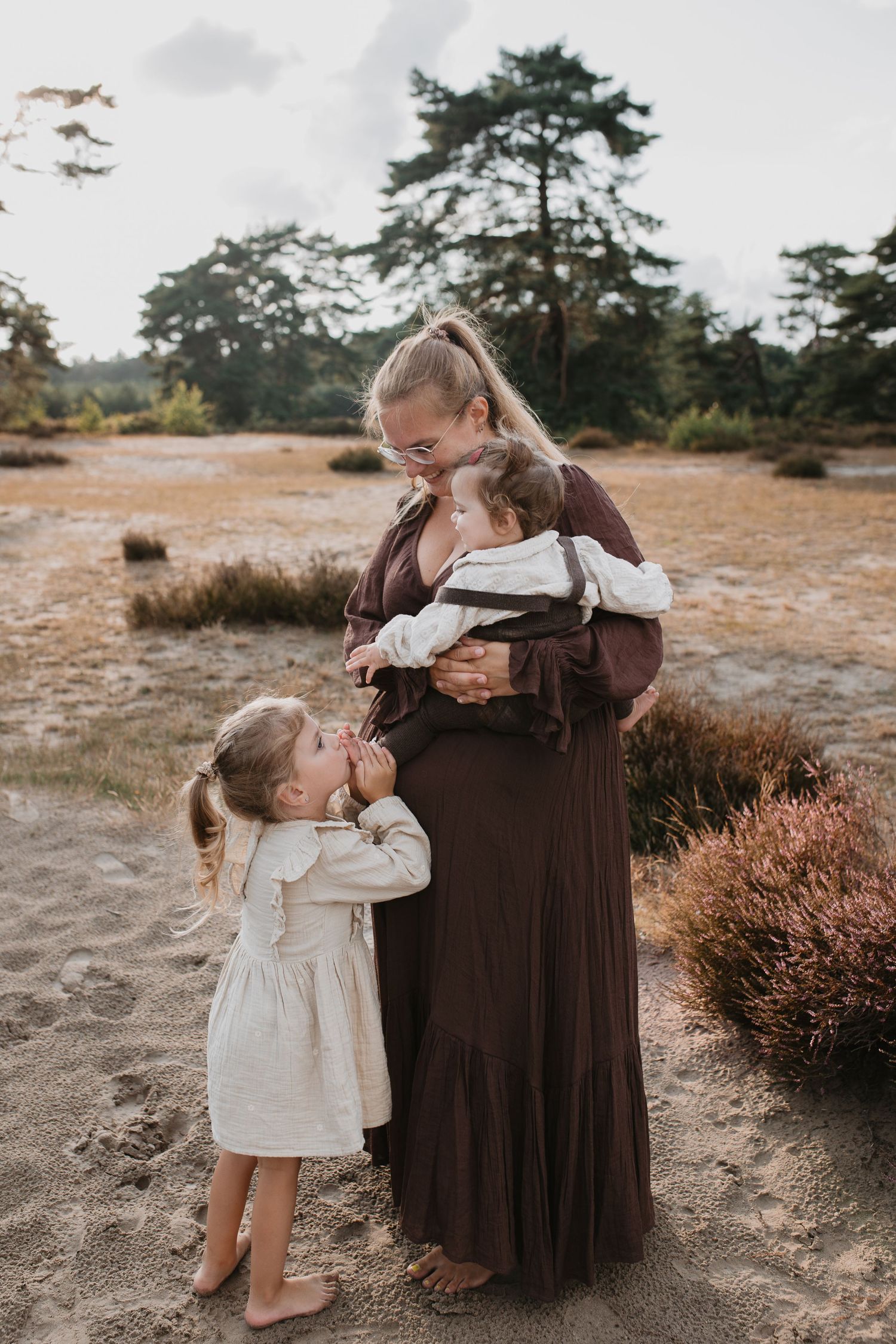 Moeder staat met haar twee dochters in een zandvlakte, de oudste kust liefdevol de voet van de baby.