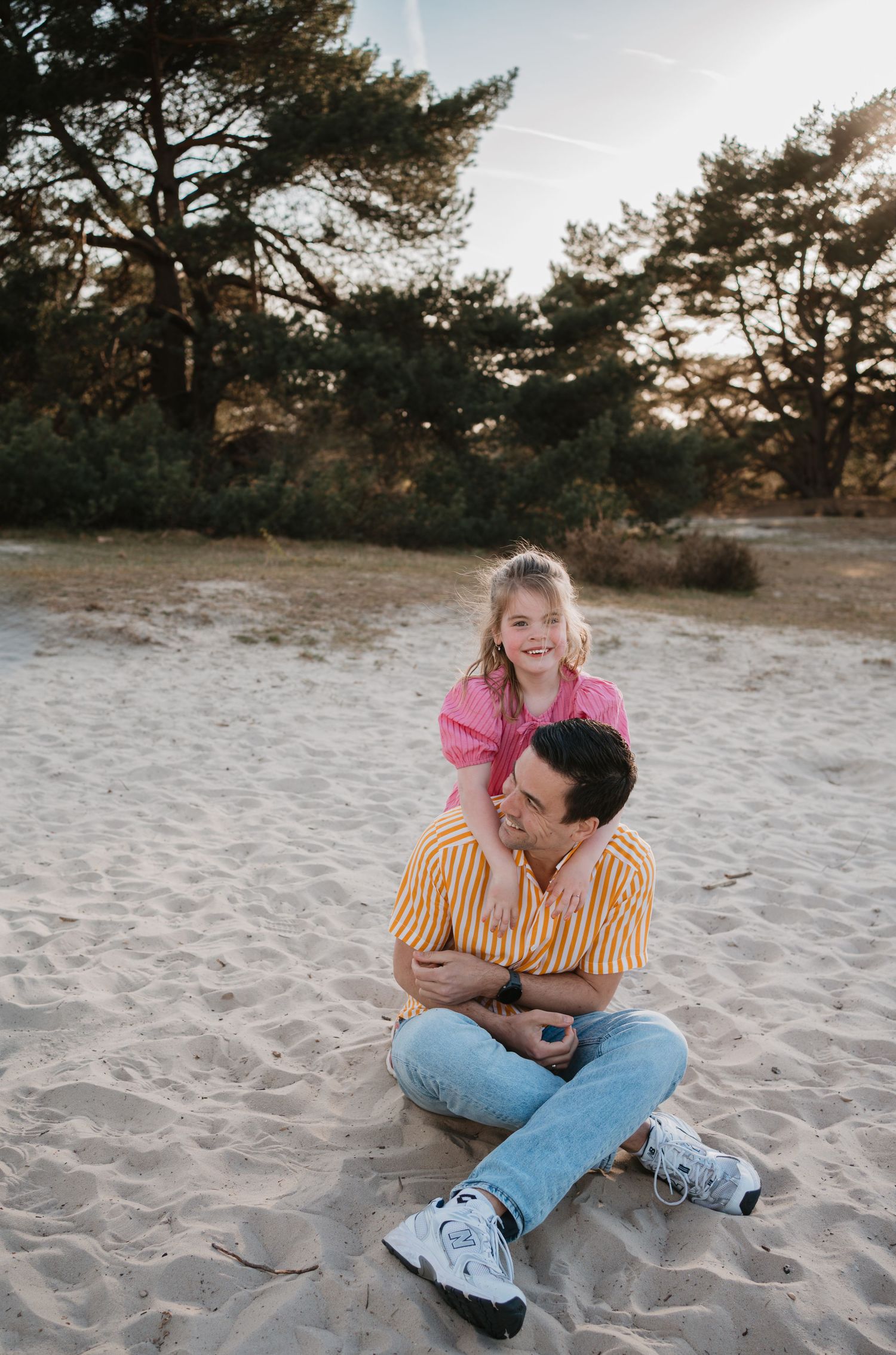 Een vader zit in het zand van de Soester Duinen en kijkt glimlachend omhoog naar zijn dochter, die liefdevol achter hem leunt.