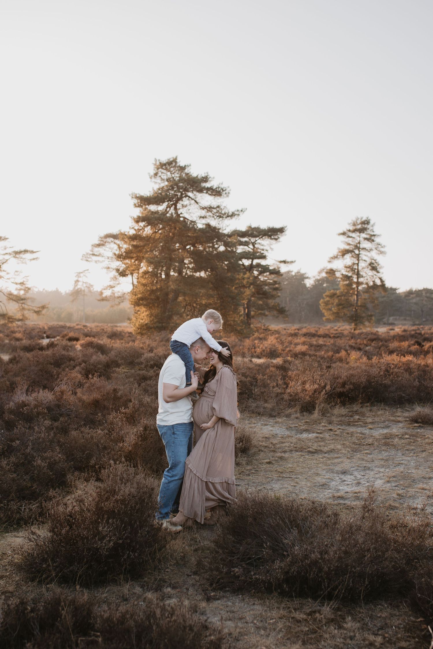 Stel, met zwangere vrouw staat met voorhoofden tegen elkaar aan. De man draagt zijn zoon op zijn schouders, die houd het hoofd van mama vast