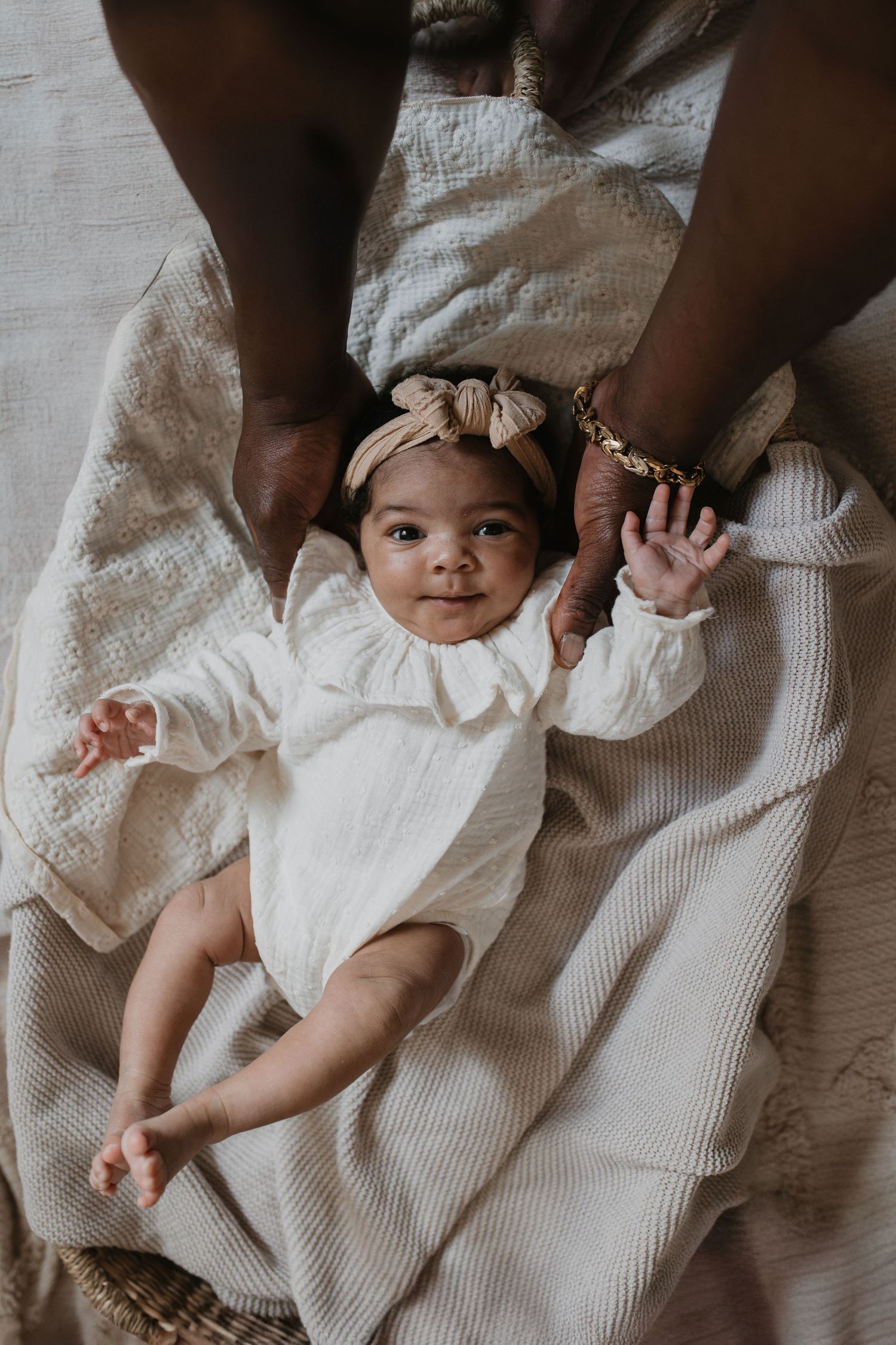 Baby met haarband kijkt lacht vanuit haar newborn mand in de camera. Zwaait naar de camera omringt door donkere handen.