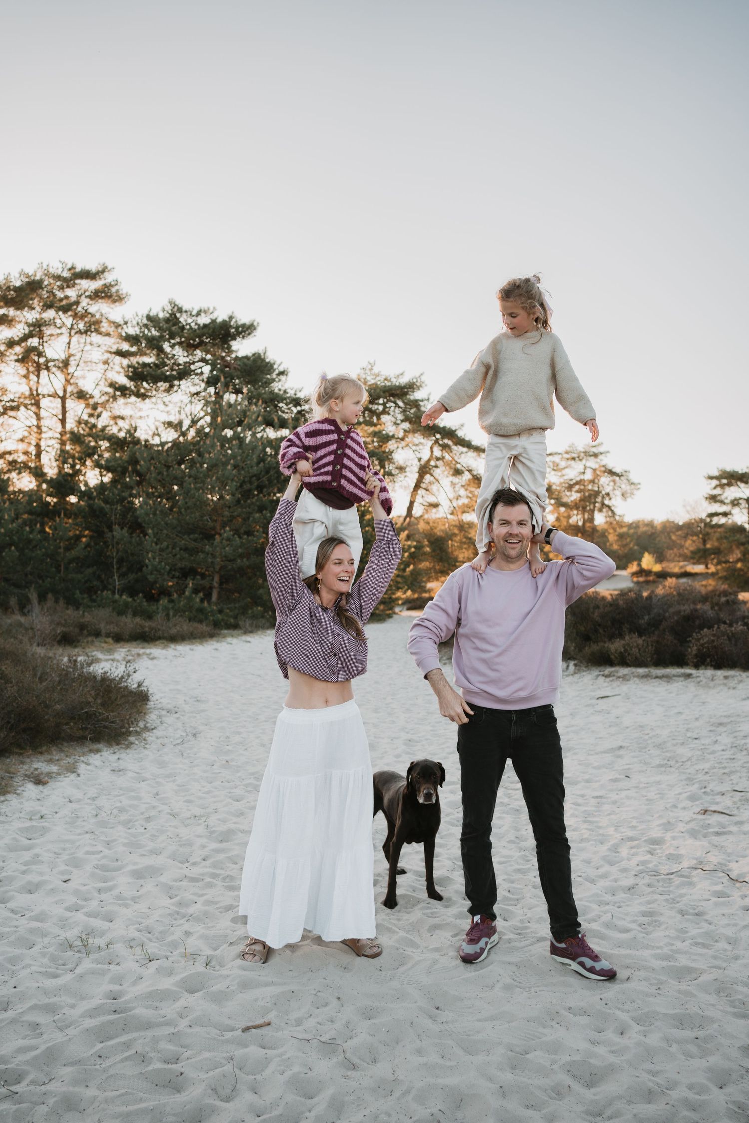 Gezin met twee jonge dochters en een hond speelt in de duinen, de ouders dragen de kinderen op hun schouders.