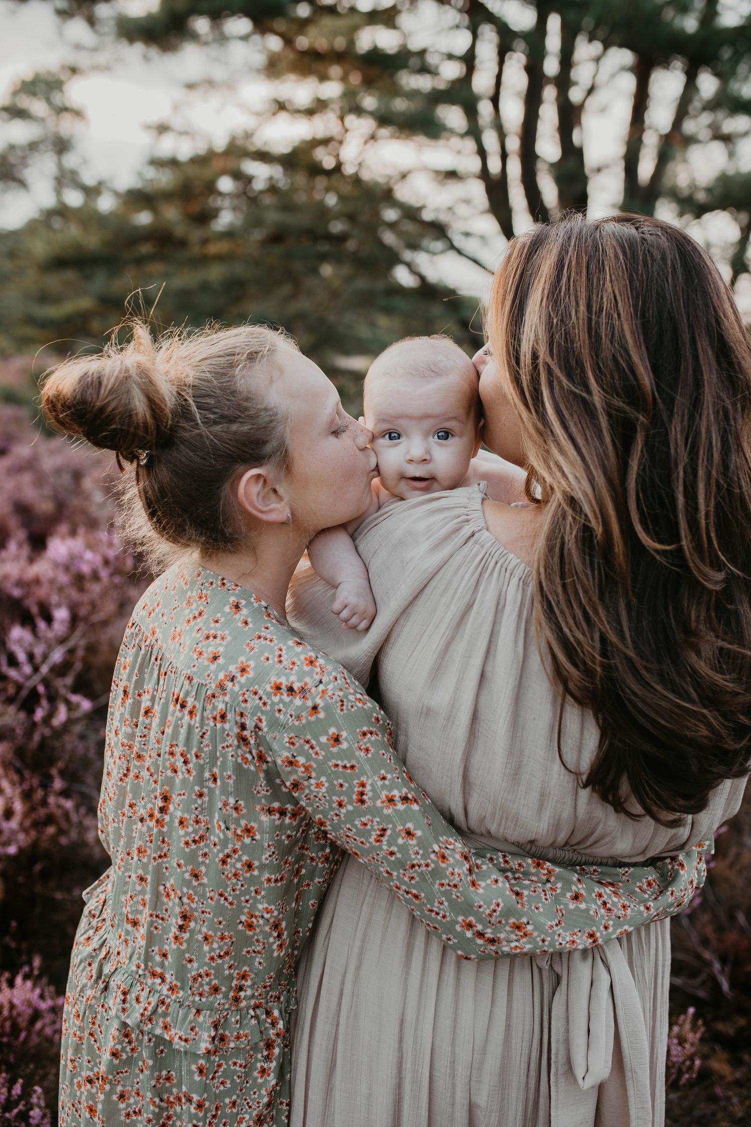 Moeder en dochter geven tegelijkertijd een kusje op de wangen van de baby, die over moeder schouder heen kijkt in de camera.