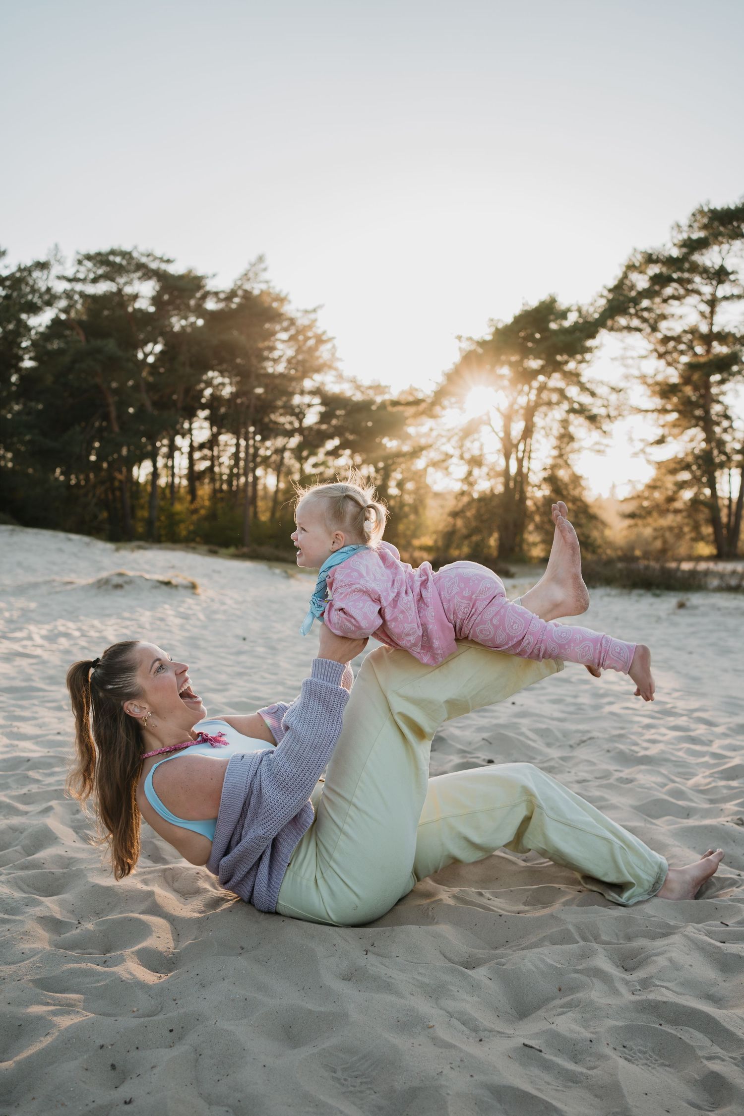 Lachende moeder balanceert haar vrolijke peuter op haar benen en handen in het zand van de Soester Duinen tijdens zonsondergang.