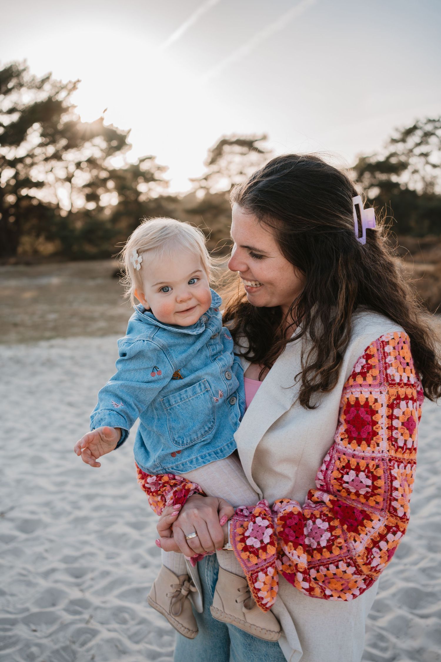 Een lachende moeder houdt haar jonge dochter liefdevol vast tijdens een fotoshoot in het zand van de Soester Duinen bij zonsondergang.