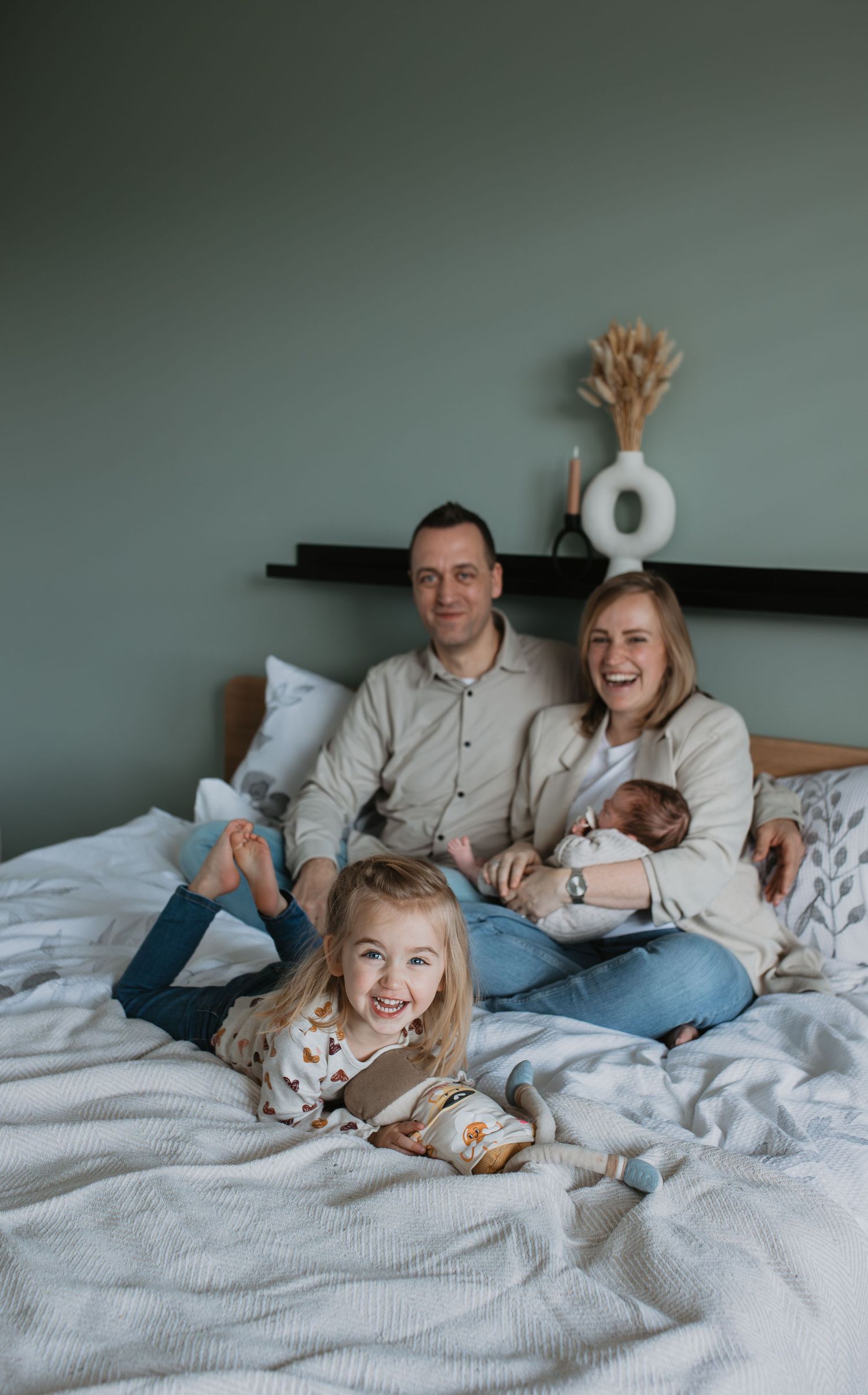 Newbornshoot met het hele gezin. Grote zus kijkt stralend in de camera terwijl de ouders met de baby op bed poseert.