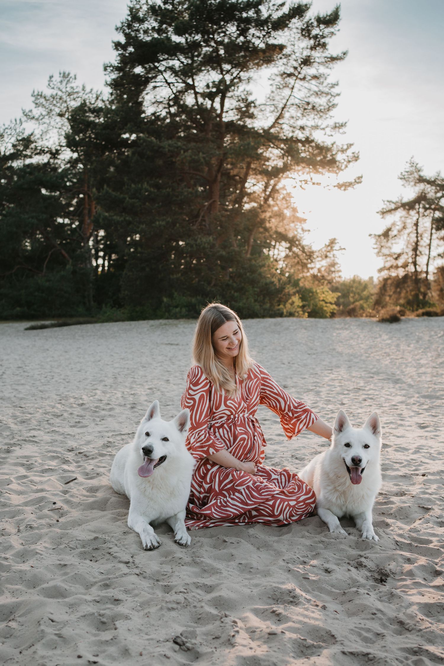 Zwangere vrouw in een rood witte jurk zit in de duinen samen met twee grote, witte honden tijdens zonsondergang.