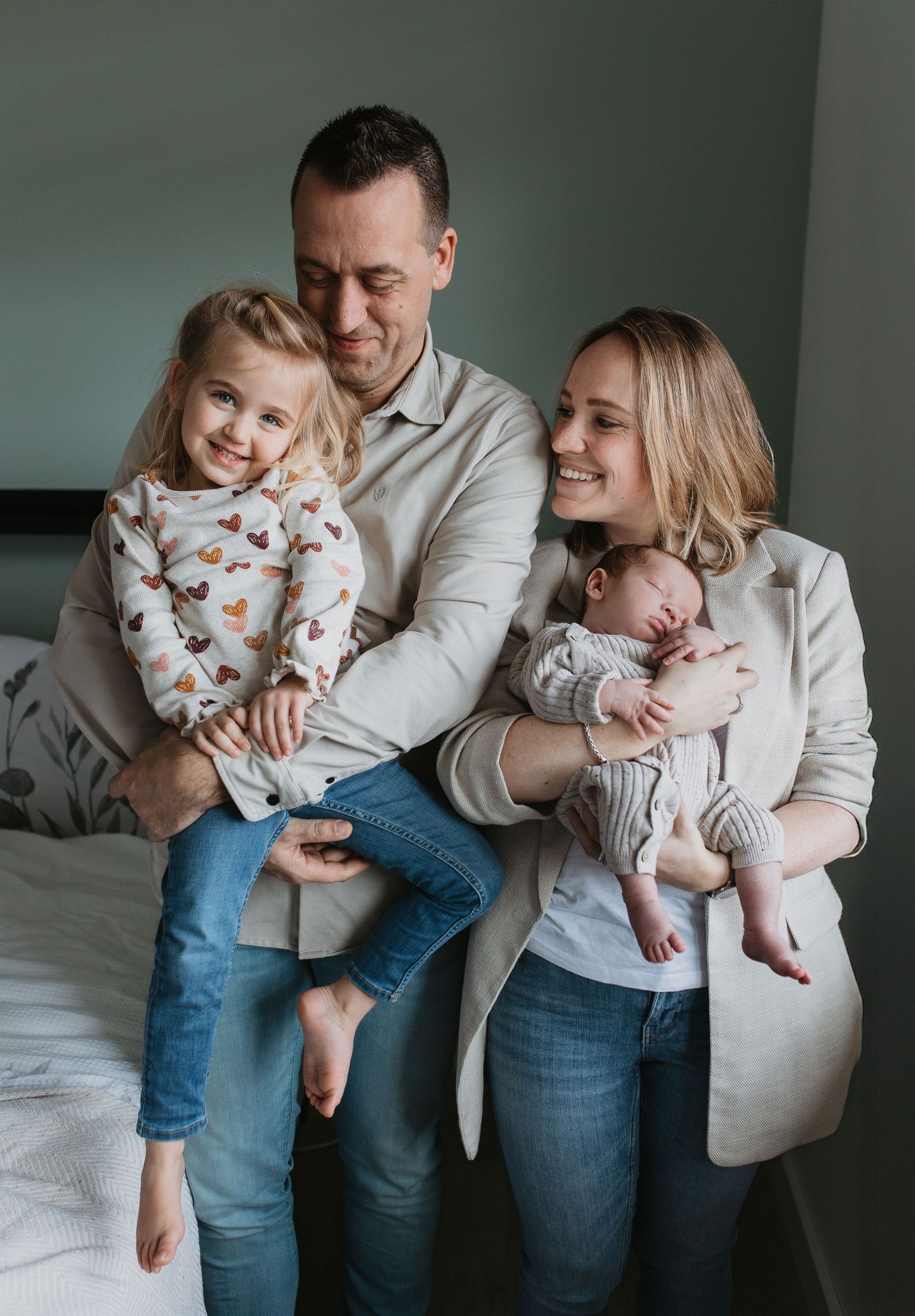Een lachende vader en moeder poseren met hun dochter en slapende newborn baby naast het bed.