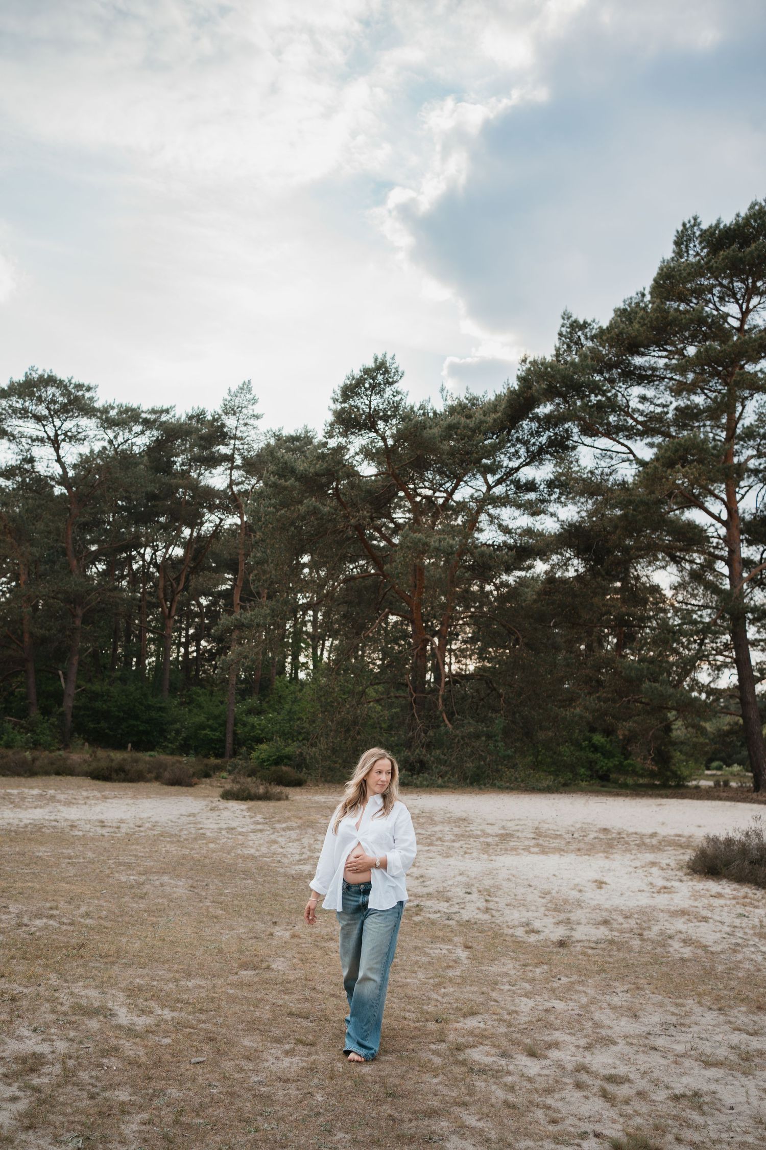 Een zwangere vrouw met los blond haar loopt in een spijkerbroek en openstaande witte blouse door een natuurgebied met bomen,de Soesterduinen