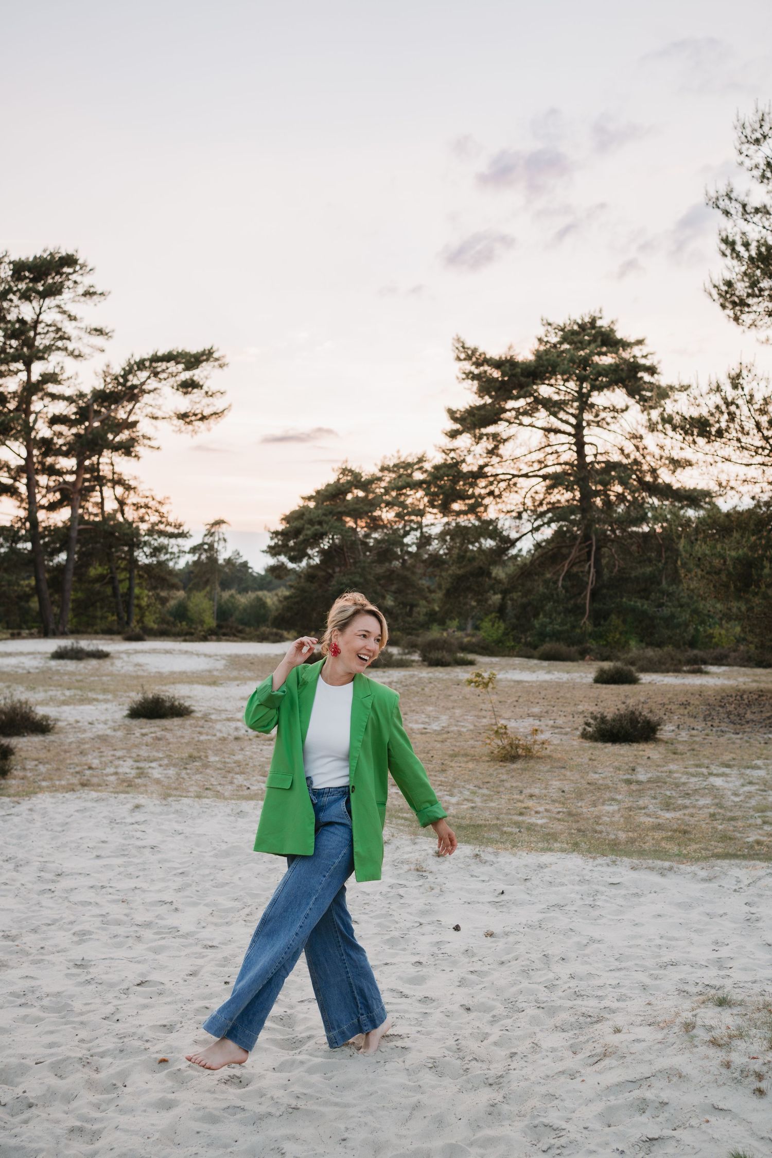 Lachende, blootsvoetse vrouw in een groene blazer en spijkerbroek wandelt over een zandvlakte, Soester Duinen tijdens de zonsondergang.